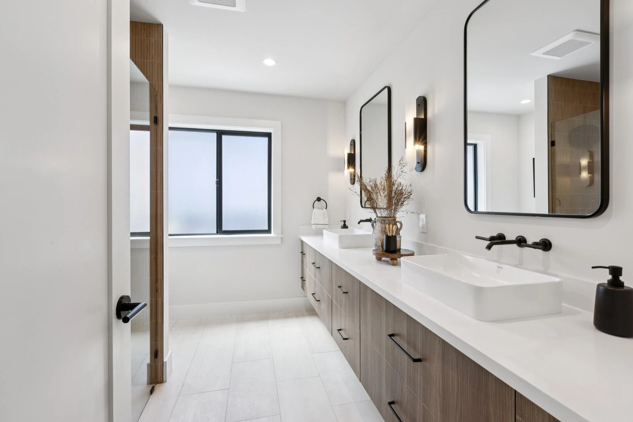 Modern bathroom with double sinks, large mirror, wooden vanity, white countertop, black fixtures, frosted window, and decorative plants.