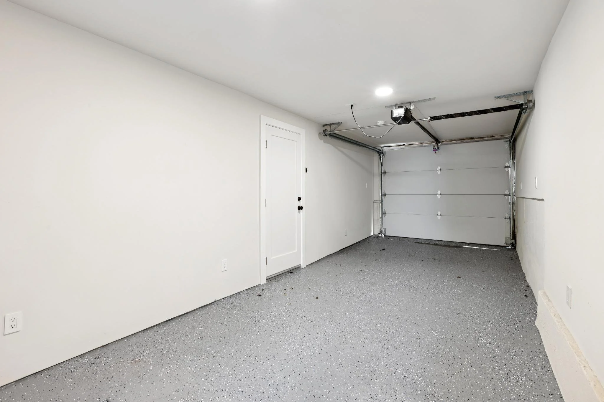 Empty garage with white walls, speckled gray epoxy flooring, a closed garage door, a white door with black doorknob, and a garage door opener mounted on the ceiling.