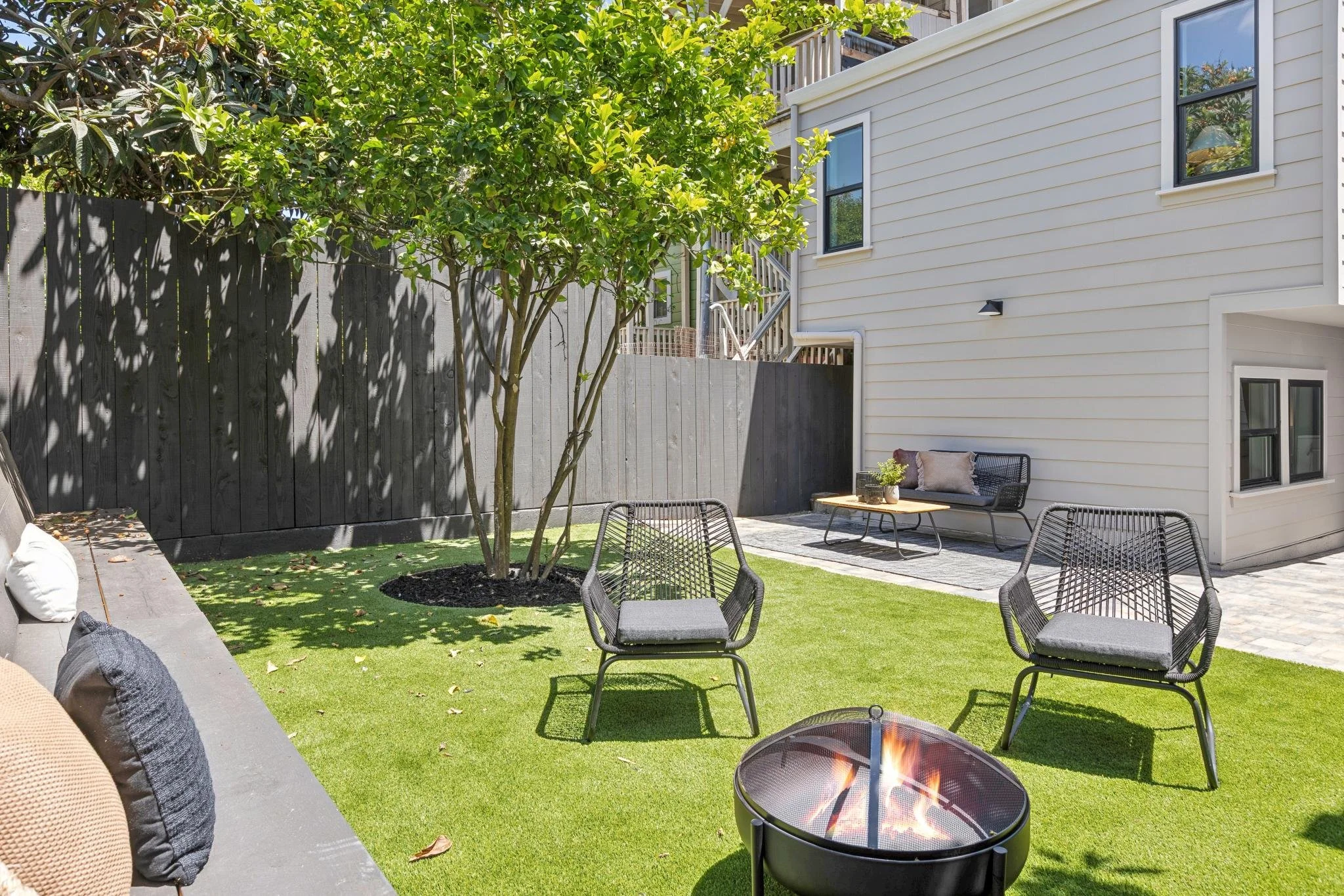 A backyard patio with outdoor furniture, a fire pit with a flame, a wooden fence, a tree, and the side of a white house with multiple windows and an outdoor light.