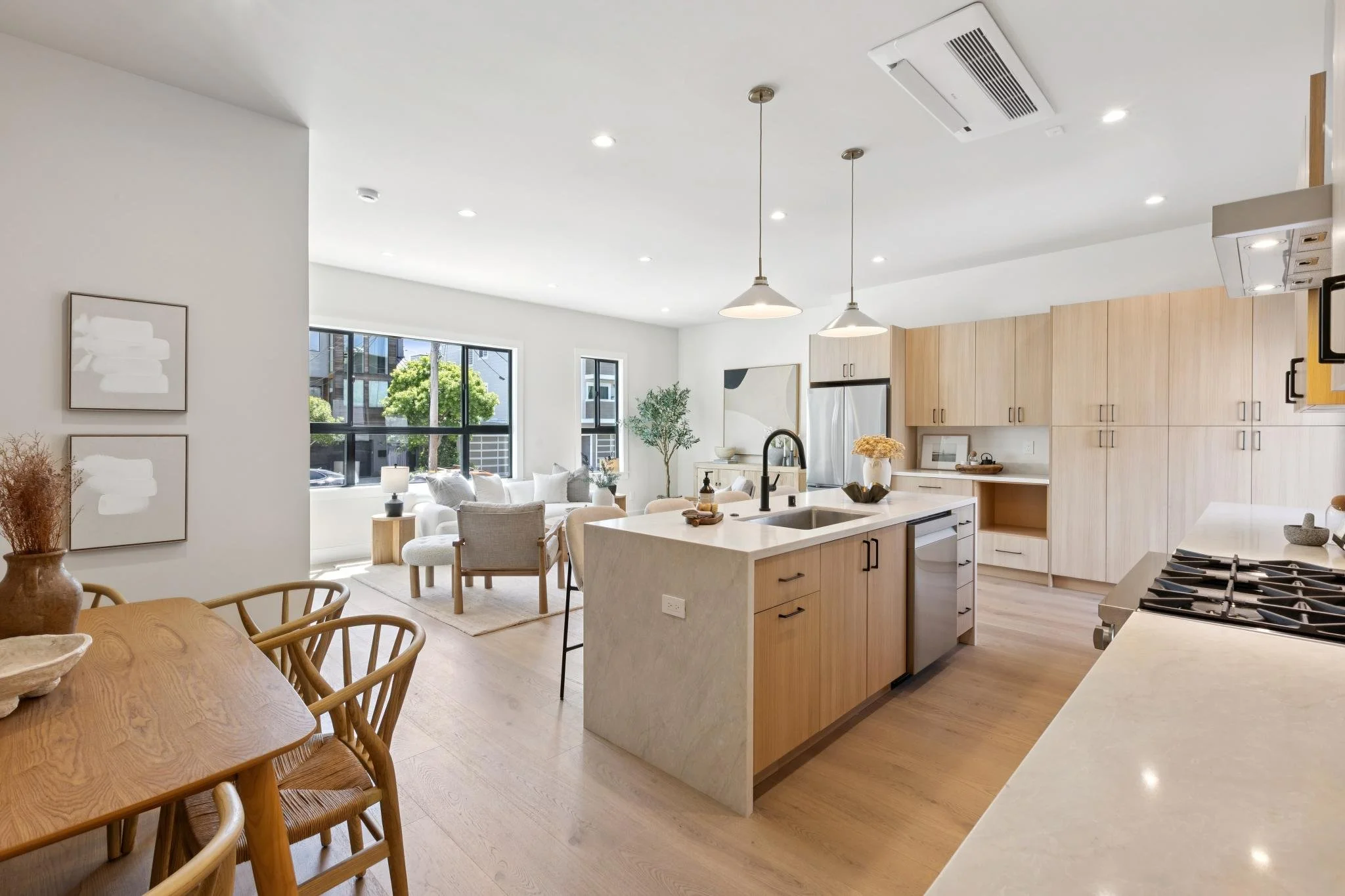 Open concept kitchen and living room with large windows and modern decor, including wooden cabinets, a kitchen island, dining table, and seating area with white and gray furniture.