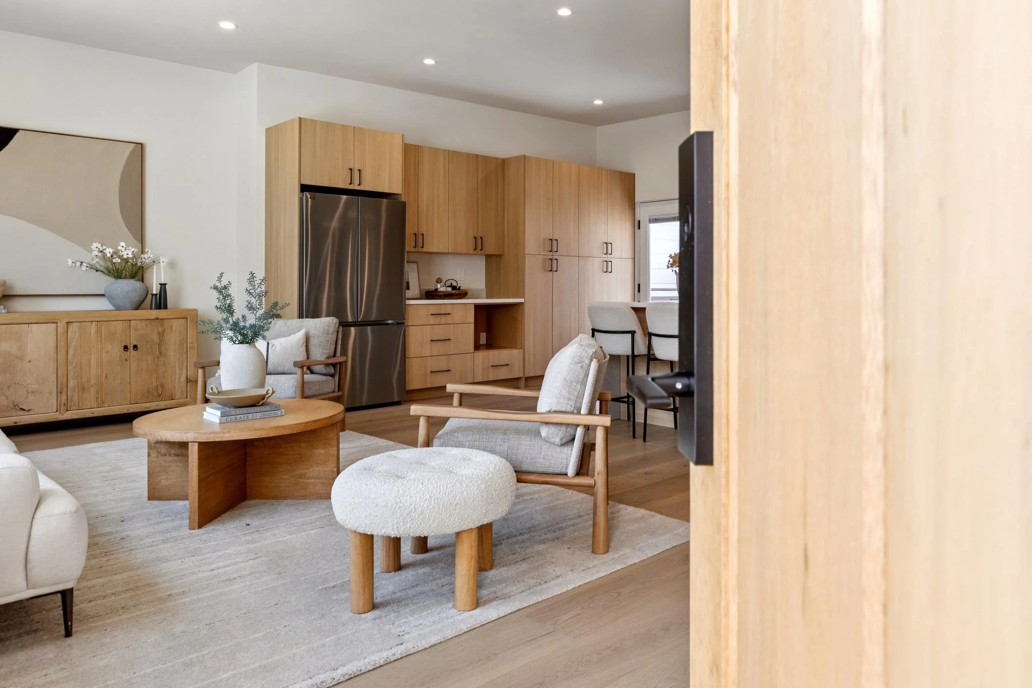 A modern living room with wooden furniture, a stainless steel refrigerator, beige armchairs, a round wooden coffee table, and decorative plants. There is a large abstract artwork on the wall and natural light from a window.