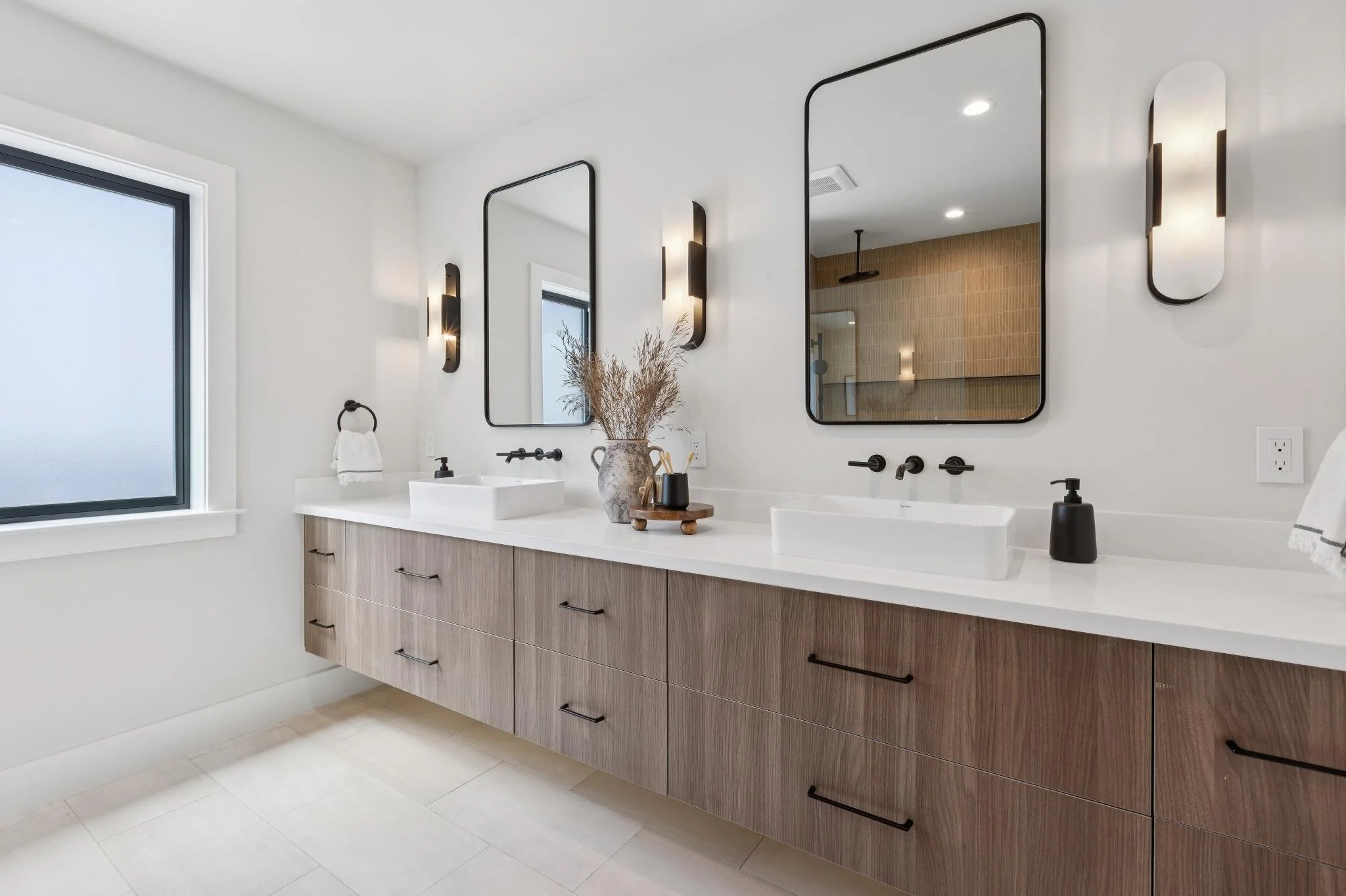 Modern bathroom with double vanity, two rectangular sinks, black fixtures, large mirrors, wooden cabinet with multiple drawers, window, and decorative vase with dried plants.