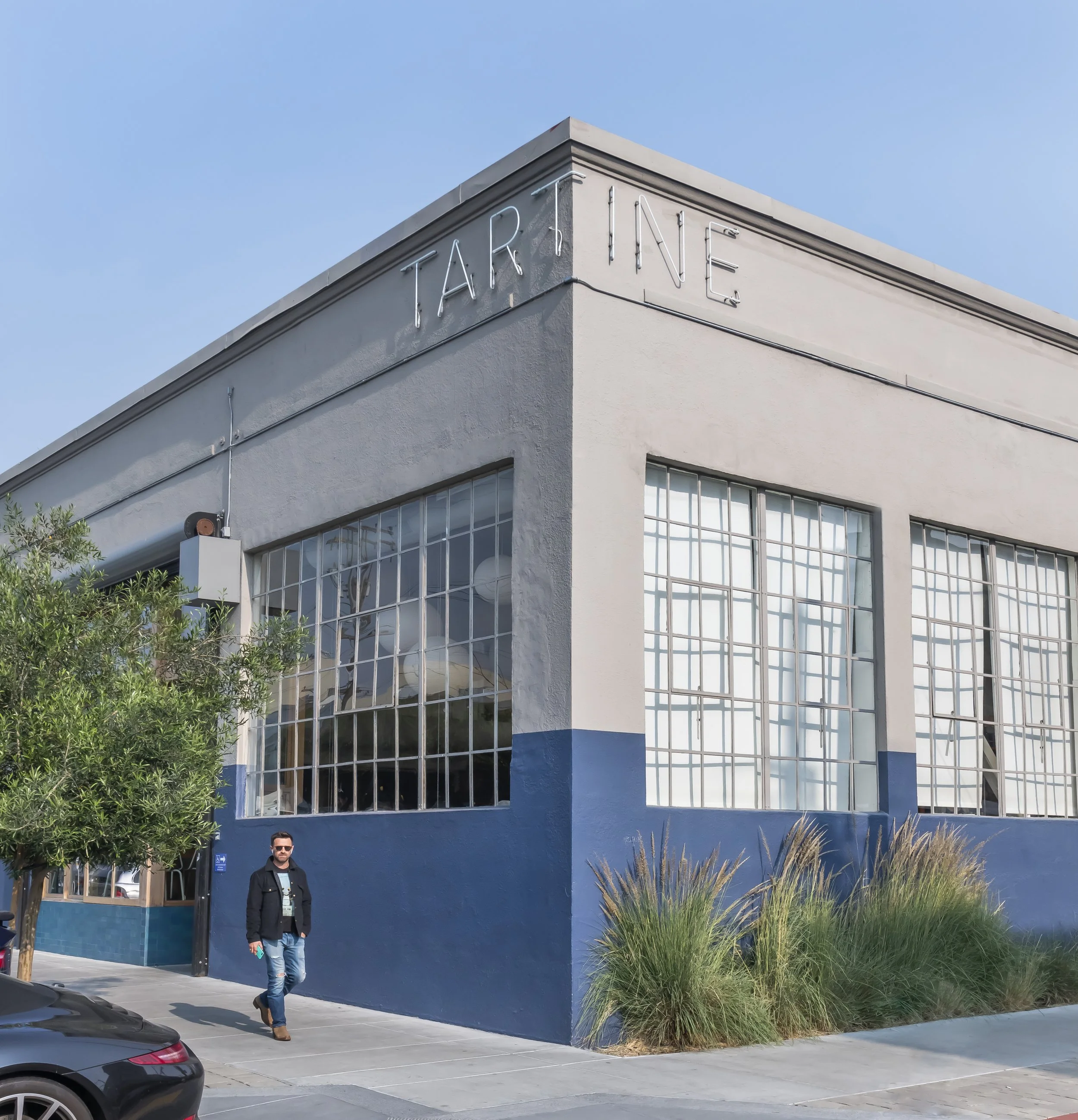 A man walking in front of a building with large windows and a neon sign that reads 'TAR INE'. The building has a modern design with a light-colored exterior, surrounding plants, and a parking lot with cars.