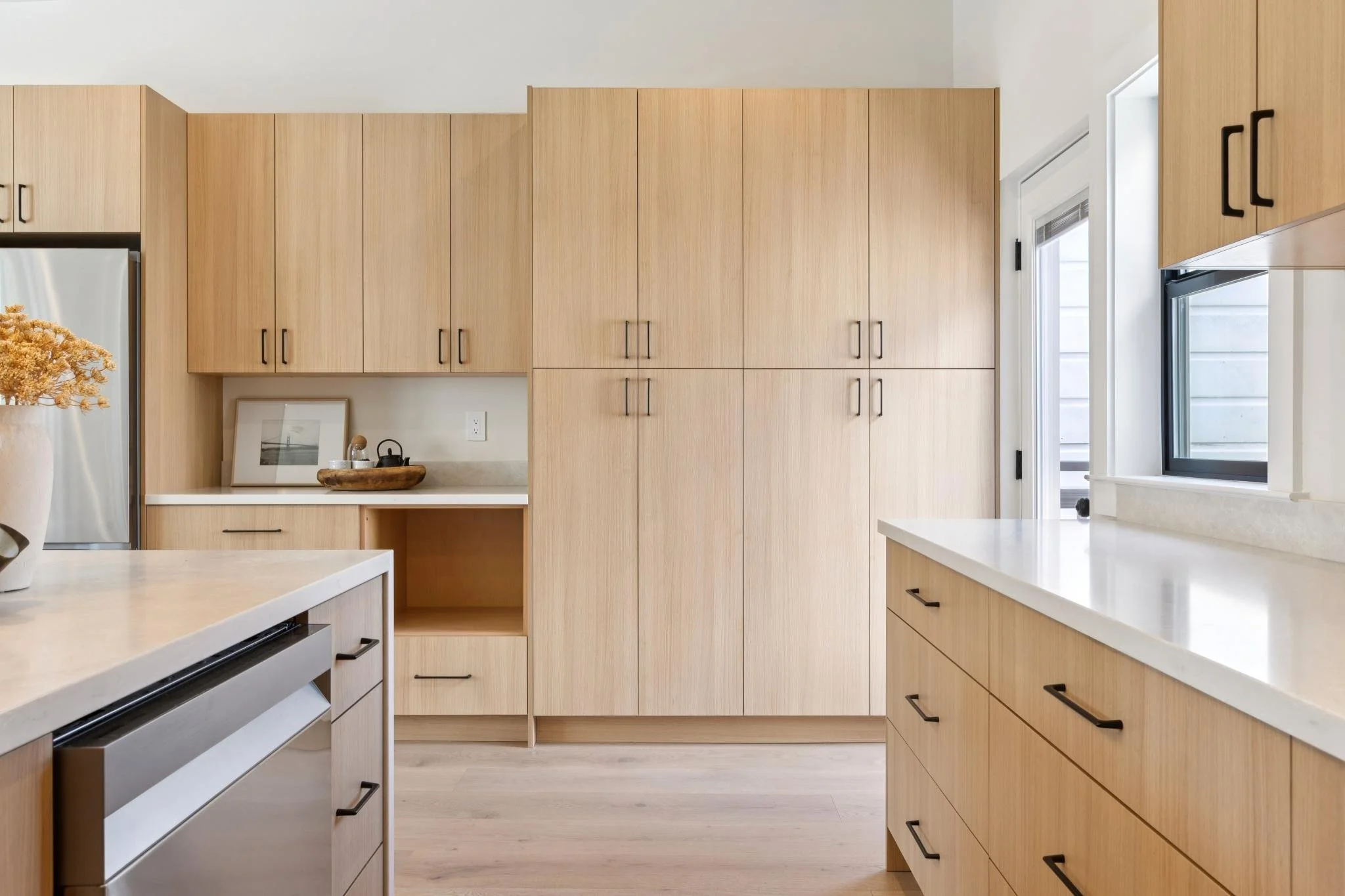 Modern kitchen with light wood cabinets, white countertops, a window, and stainless steel appliances.