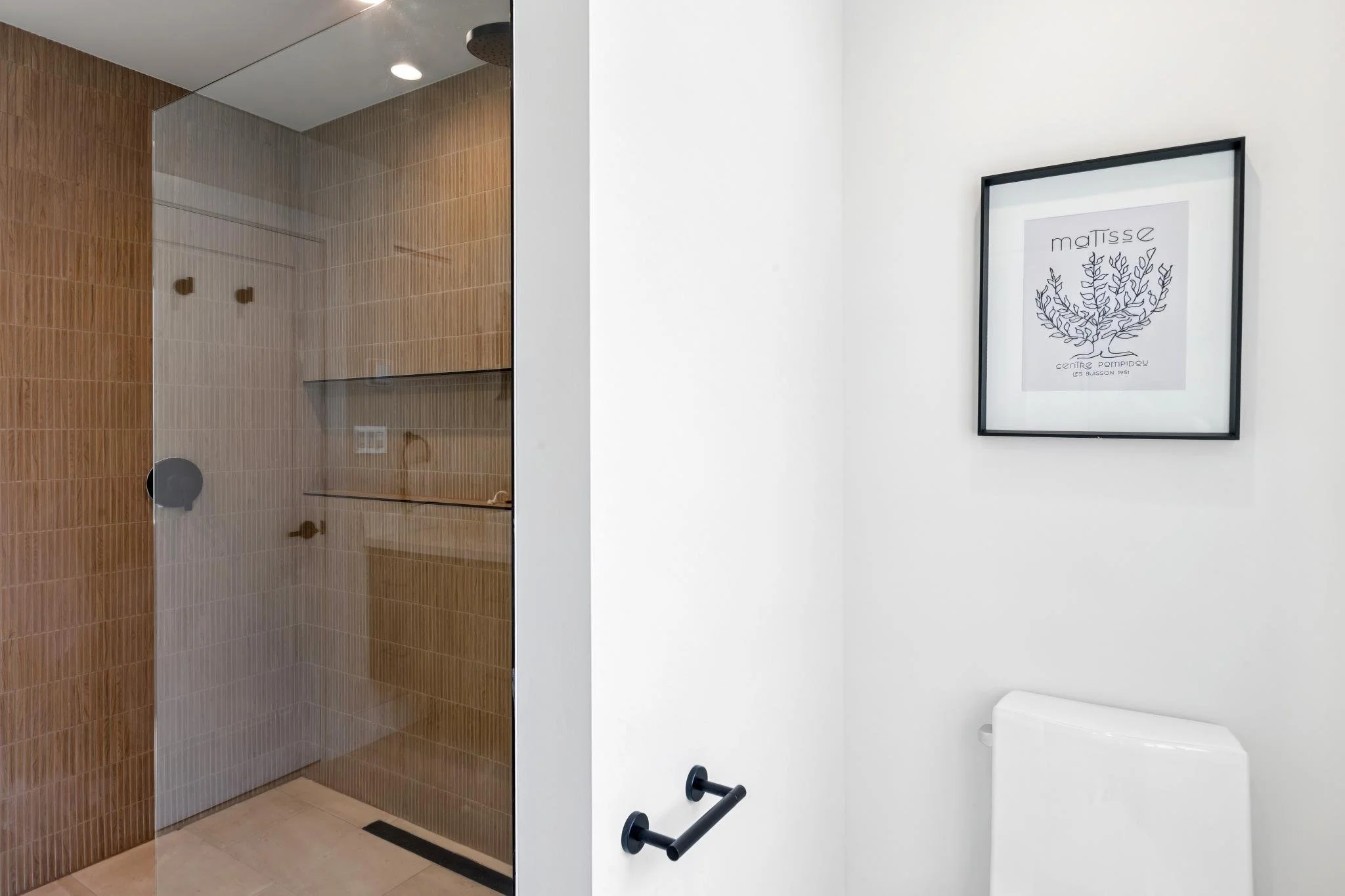 A modern bathroom with a frosted glass shower enclosure with brown tiles on the wall inside the shower. To the right, there is a white wall with a framed black and white artwork featuring botanical line drawings and text. A toilet paper holder and pa