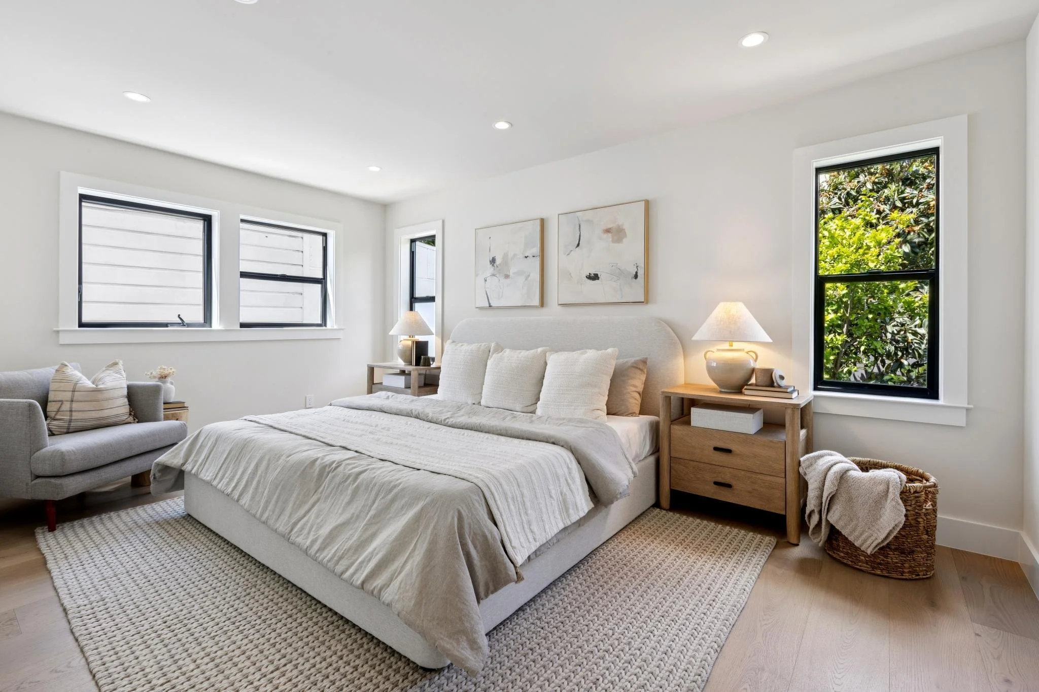 A bright and airy bedroom with white walls and ceiling, featuring three windows with black frames, a bed with white bedding and pillows, two wooden bedside tables with lamps, and modern abstract art on the wall. There is a small sofa with pillows and a rug on the light wood floor, with a wicker basket and blanket nearby.
