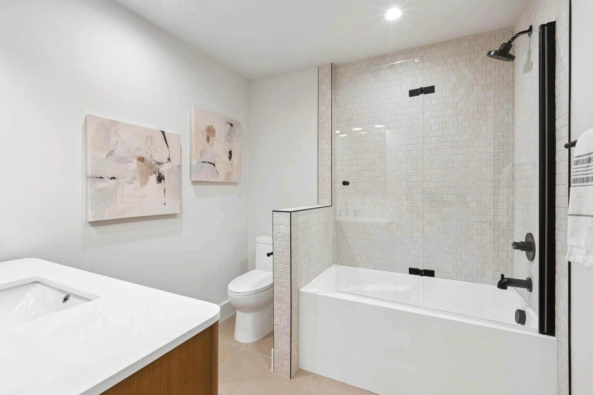 Modern bathroom with a white bathtub, a toilet, a shower with glass doors and black fixtures, and abstract artwork on the wall.