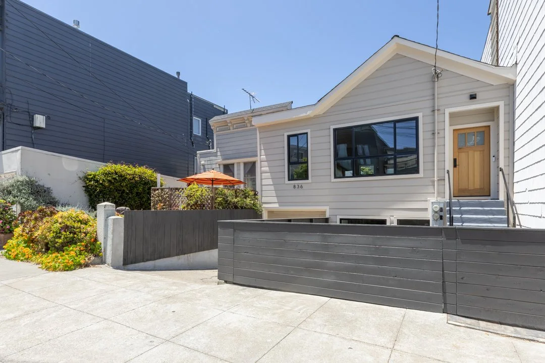 Front view of a light-colored house with large windows, a wooden front door, stairs, and a small garden with bushes and an orange umbrella.