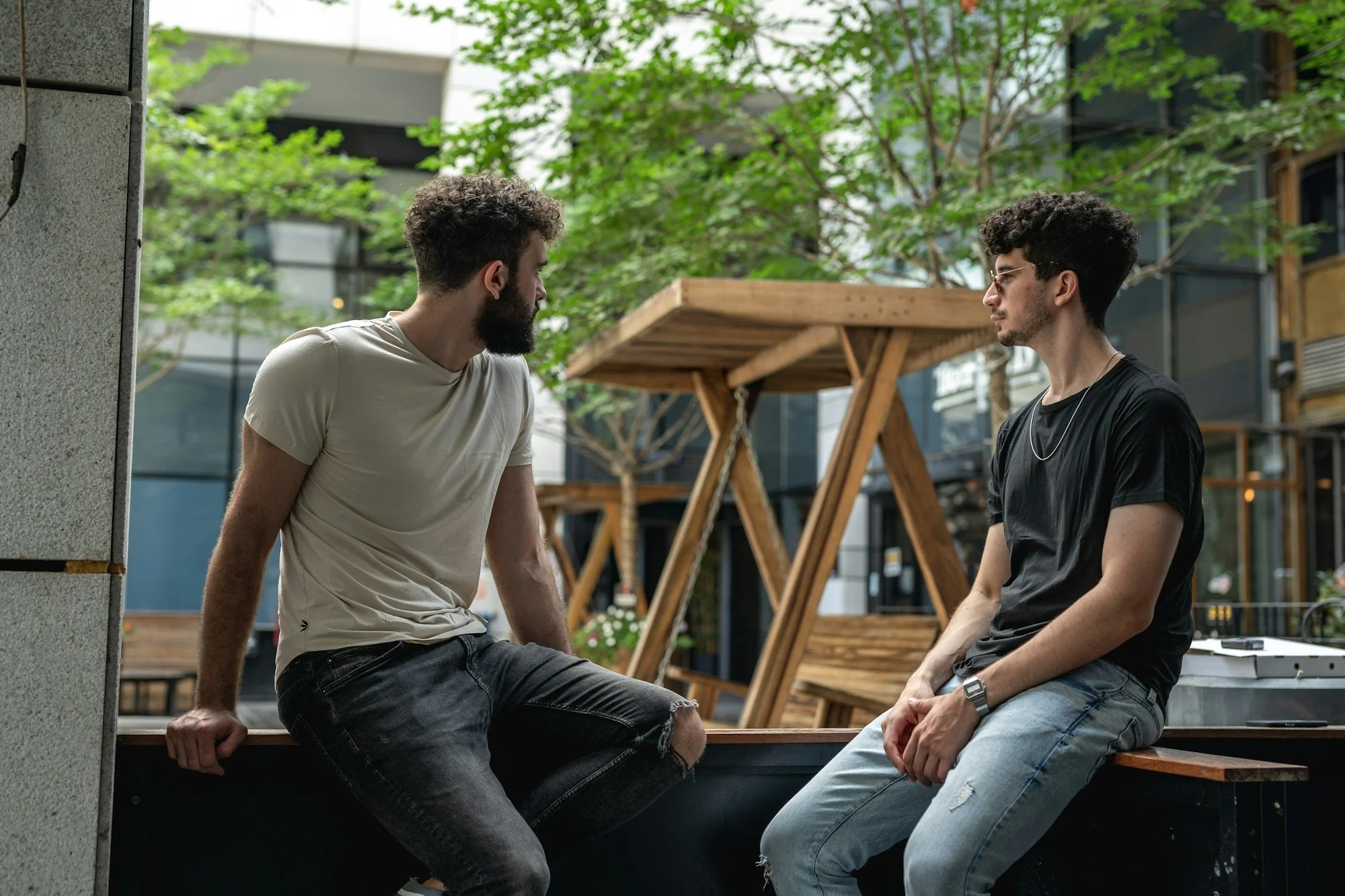 Two men sit facing each other on a wooden bench in an outdoor courtyard with trees.