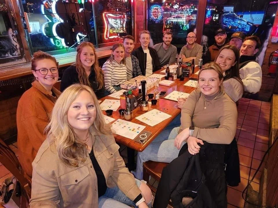 A group of fifteen people seated around a long wooden table in a restaurant, smiling at the camera, with neon signs visible in the background.