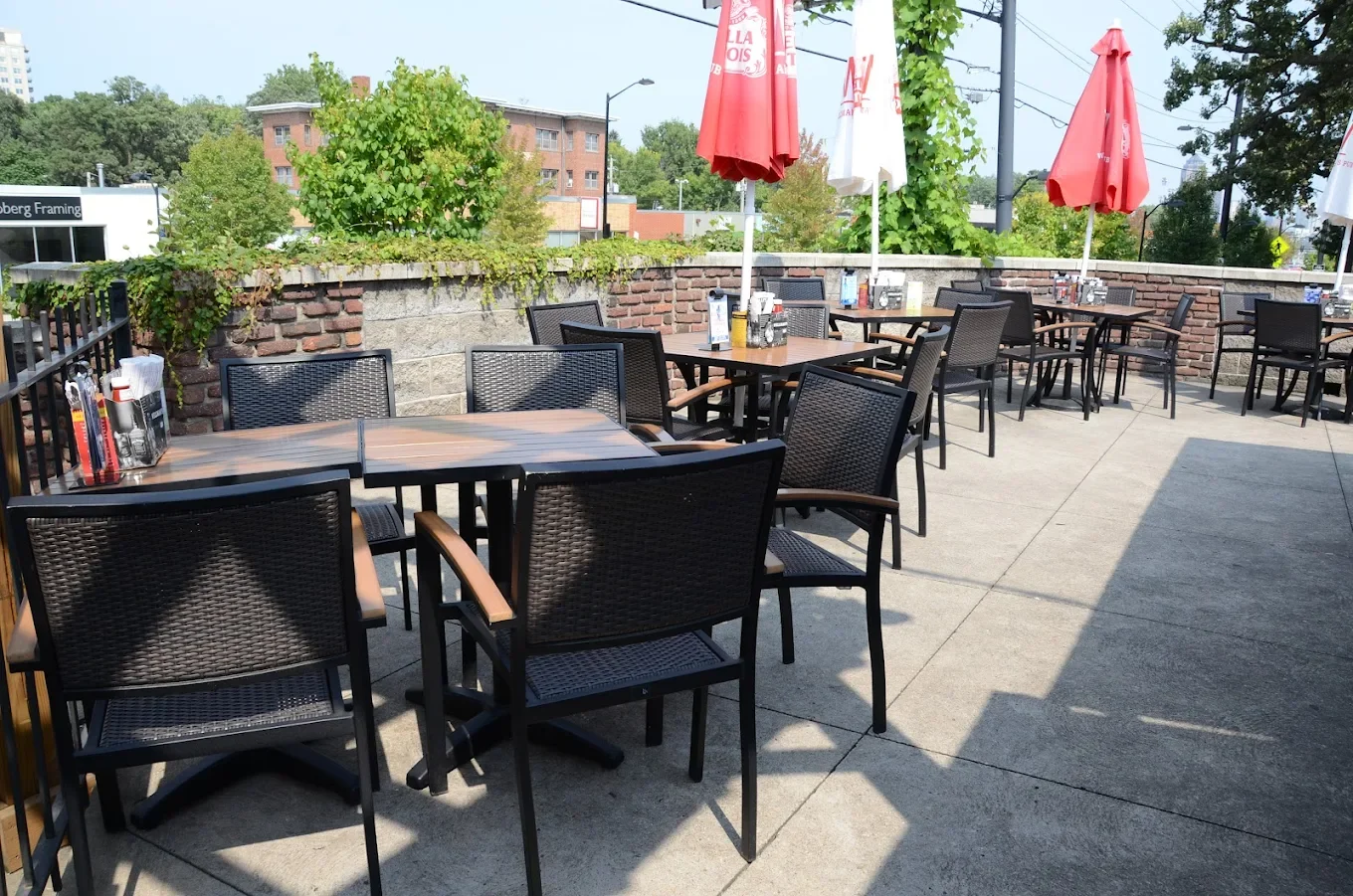 Empty outdoor patio with tables, black chairs, red and white umbrellas, and a brick wall, with trees and buildings in the background.