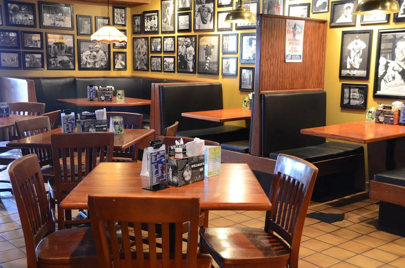 Empty restaurant with wooden tables, chairs, and black booths, decorated with black and white photographs on yellow walls, with pendant lights hanging from the ceiling.