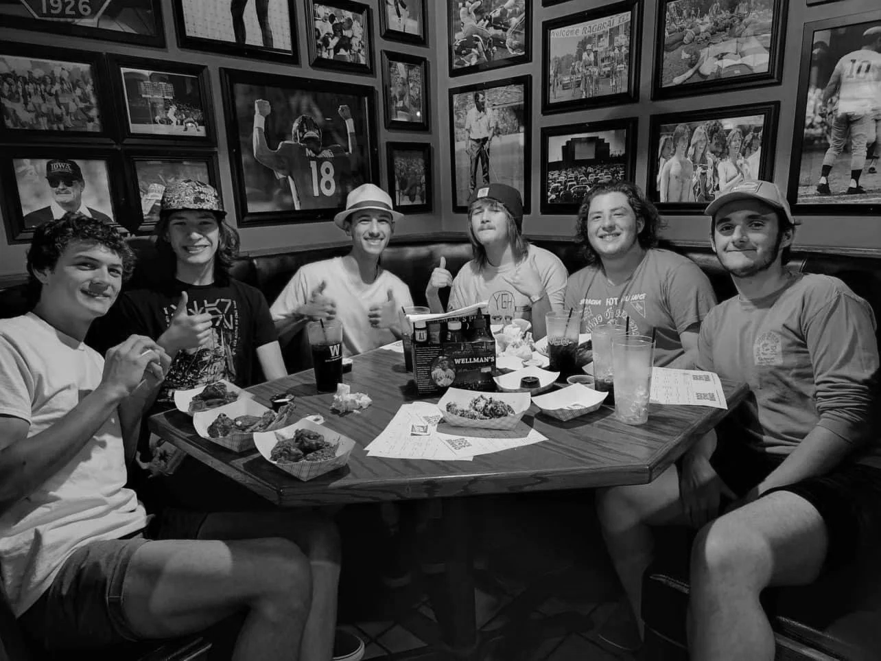 Group of six young adults sitting at a restaurant table with food and drinks, smiling and enjoying their time, surrounded by sports-themed photos on the wall in black and white.