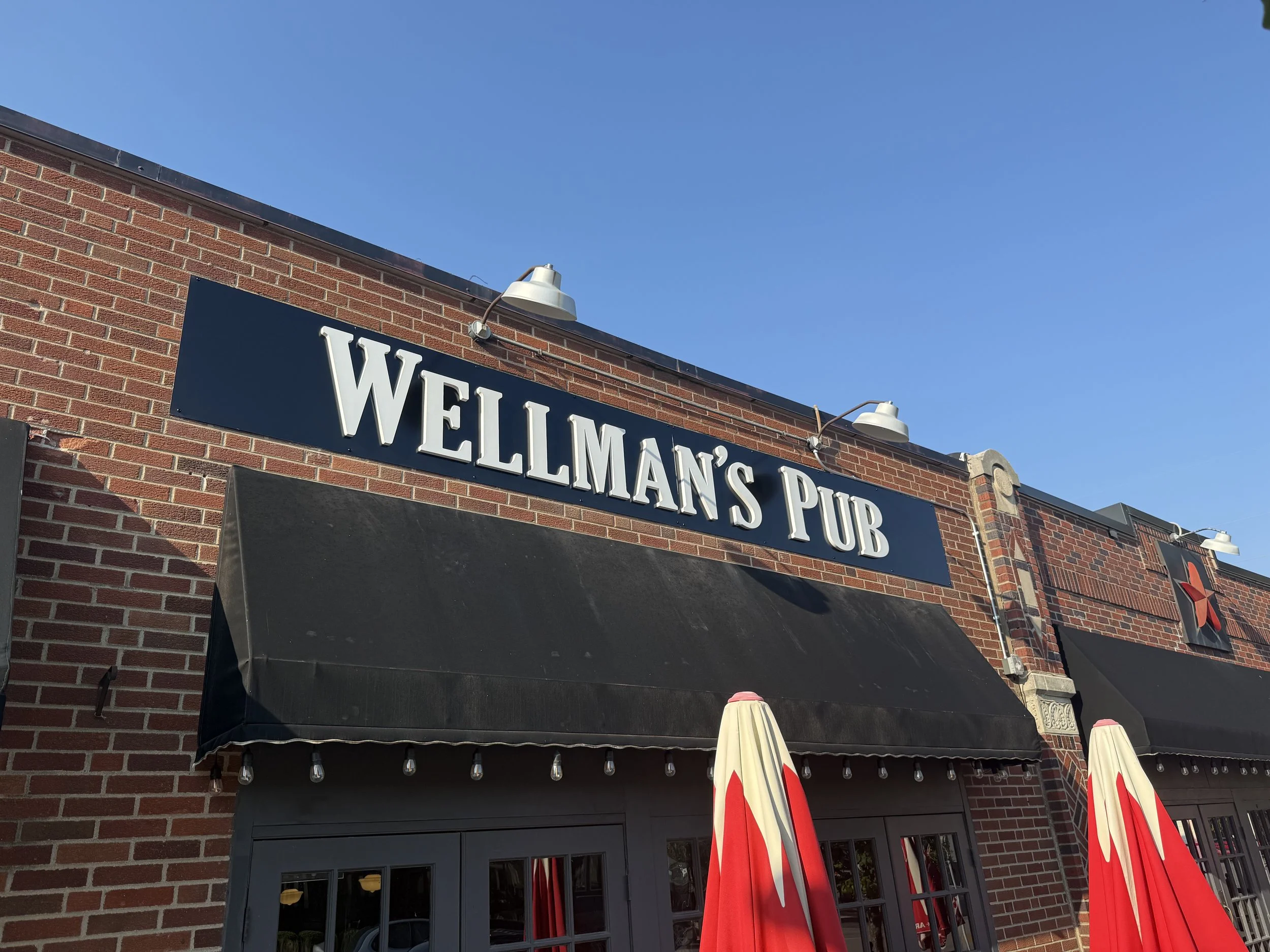 Exterior of a brick building with a sign that reads 'WELLMAN'S PUB', two outdoor lamps, black awning, and red and white umbrellas.