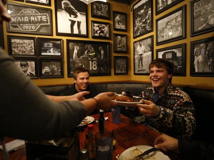 Two smiling young men in a restaurant receiving a birthday cake from a server. The restaurant has vintage black-and-white framed photos on the yellow walls.