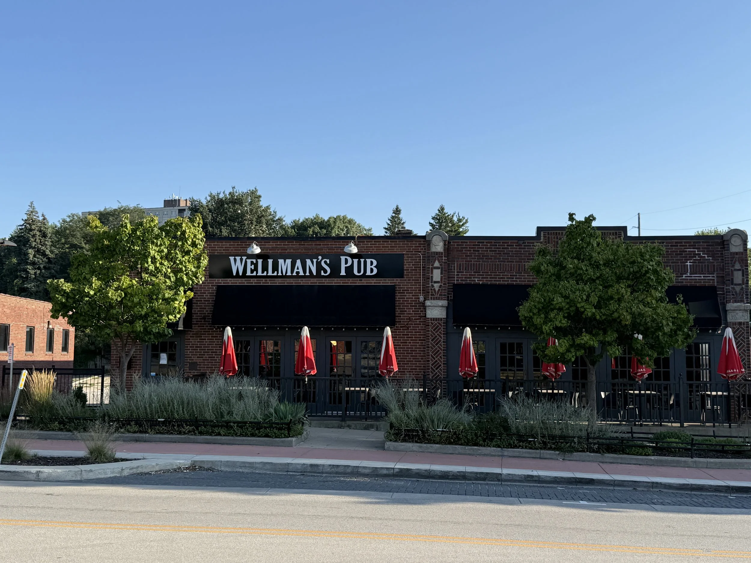 Exterior of Wellman's Pub with red brick facade, black sign, patio umbrellas, and lush green trees against a blue sky.