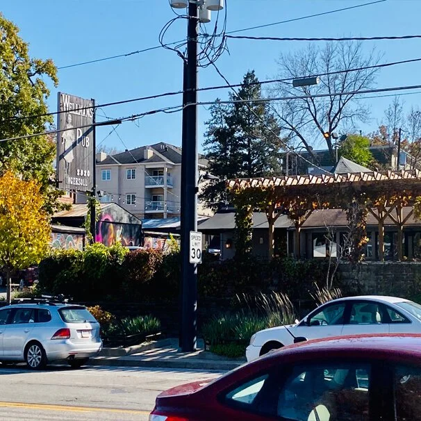 Street scene with parked cars, utility pole with wires, a building with painted murals, and a house with a roof vine trellis, under a clear sky.