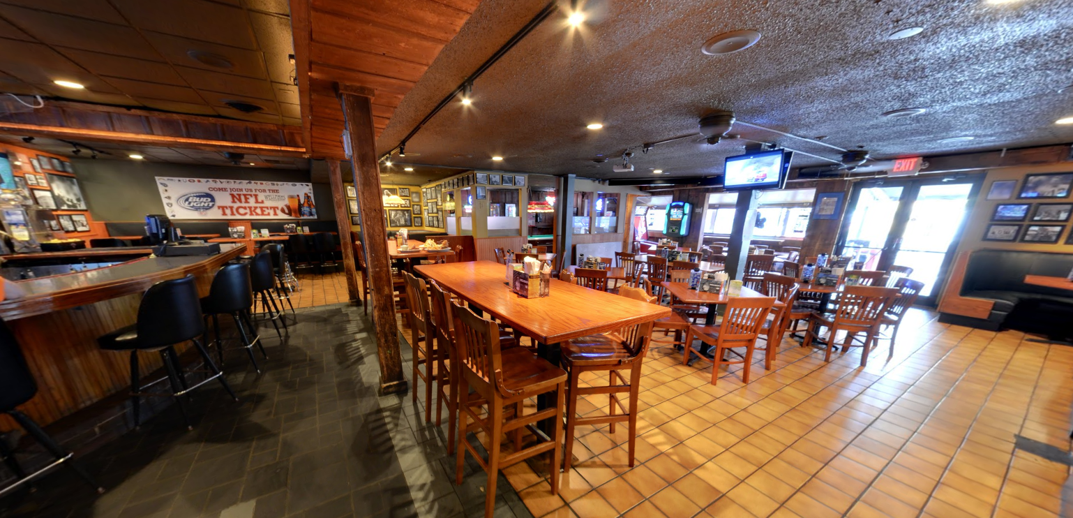 Interior of a restaurant or bar with wooden tables and chairs, a ceiling-mounted TV, framed pictures on the walls, and large windows at the front with an exit door.