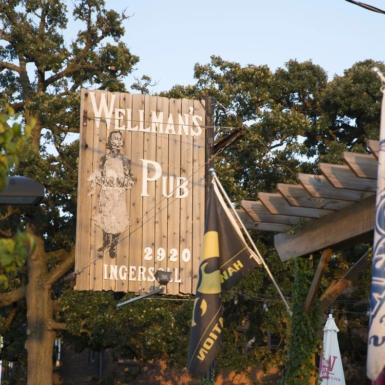 Wooden sign for Wellman's Pub with an illustration of a woman, located at 2920 Ingersoll. Flags are hanging nearby, and trees are in the background.