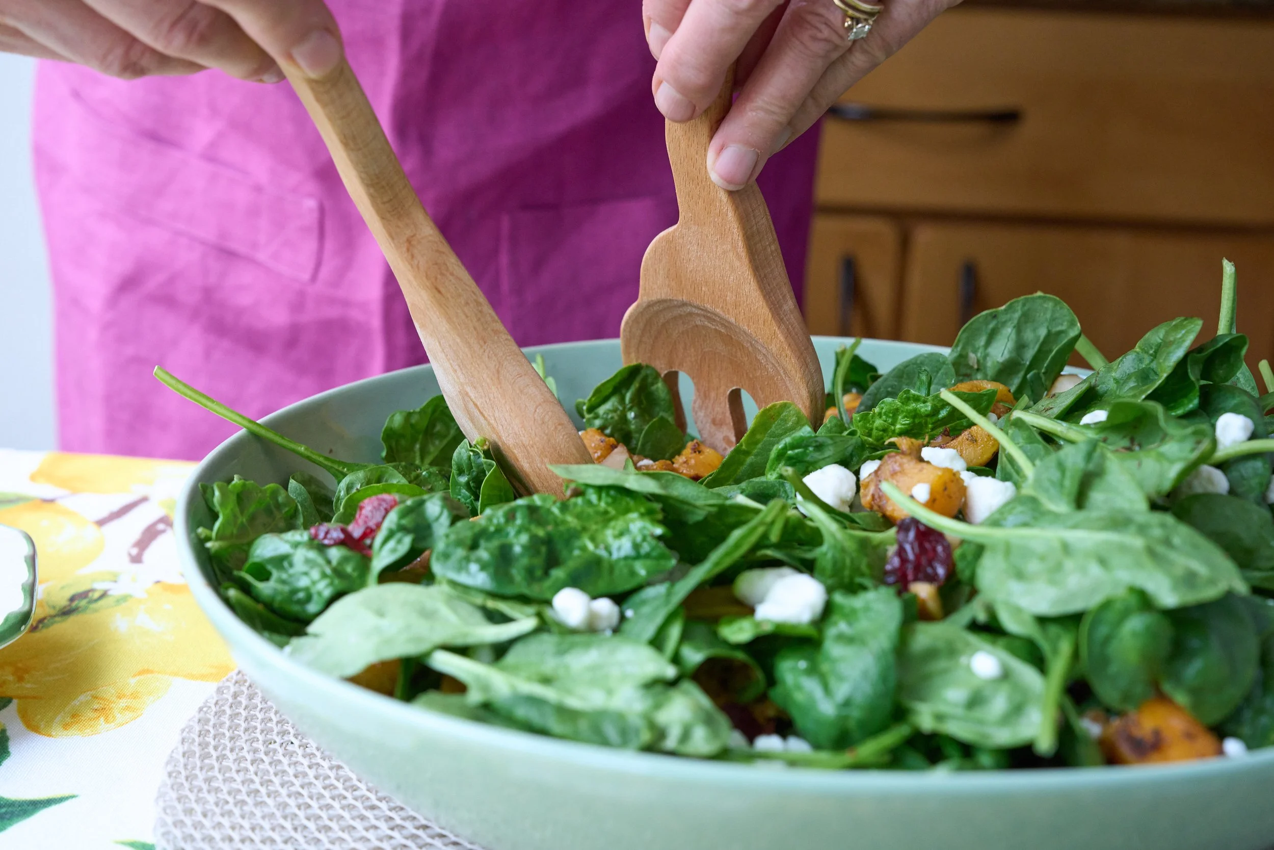 Close-up of a bowl filled with mixed greens, colorful vegetables, and cheese, with two wooden salad tongs in use, on a table with a yellow floral tablecloth.