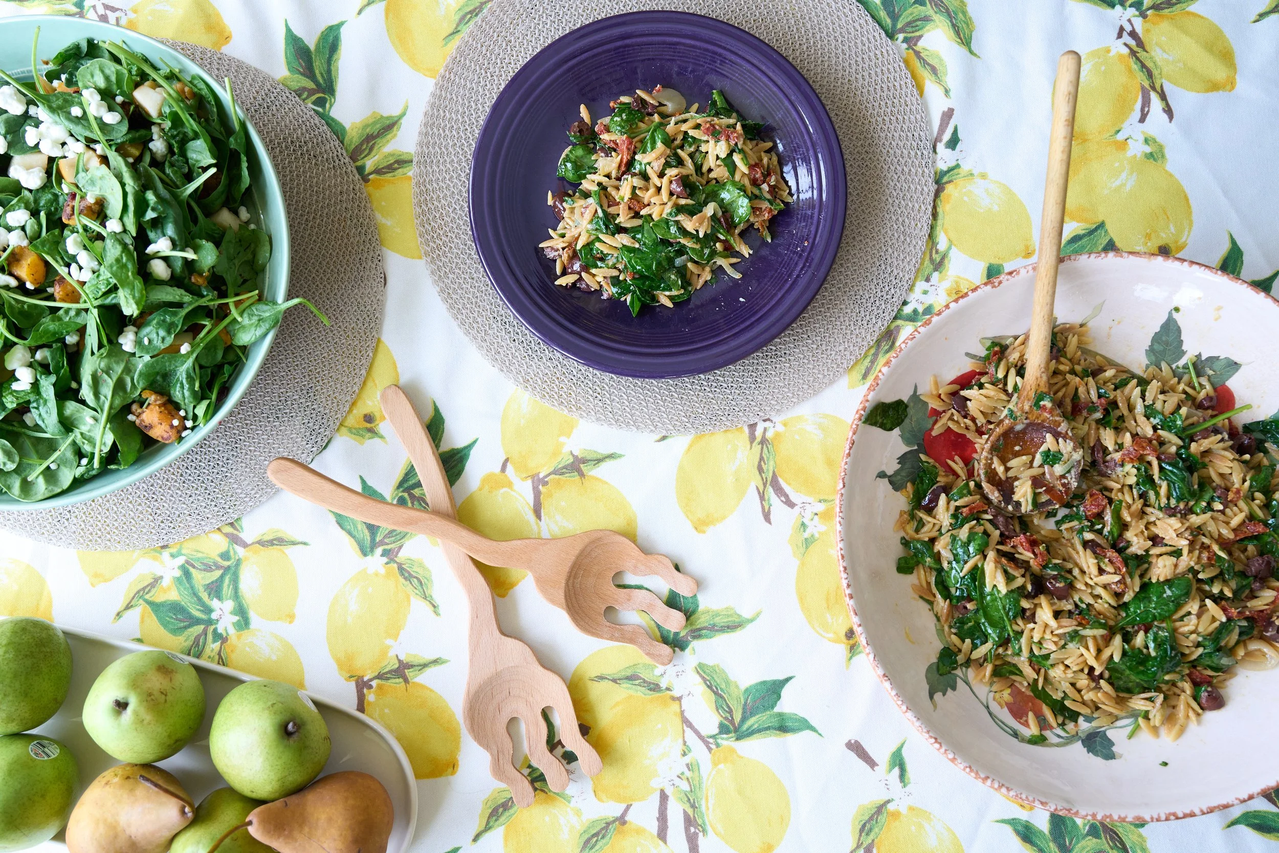 A table set with pasta salad on a purple plate, a green salad, a bowl with more pasta salad, a bowl of green apples and pears, and wooden salad servers, all on a lemon-patterned tablecloth.