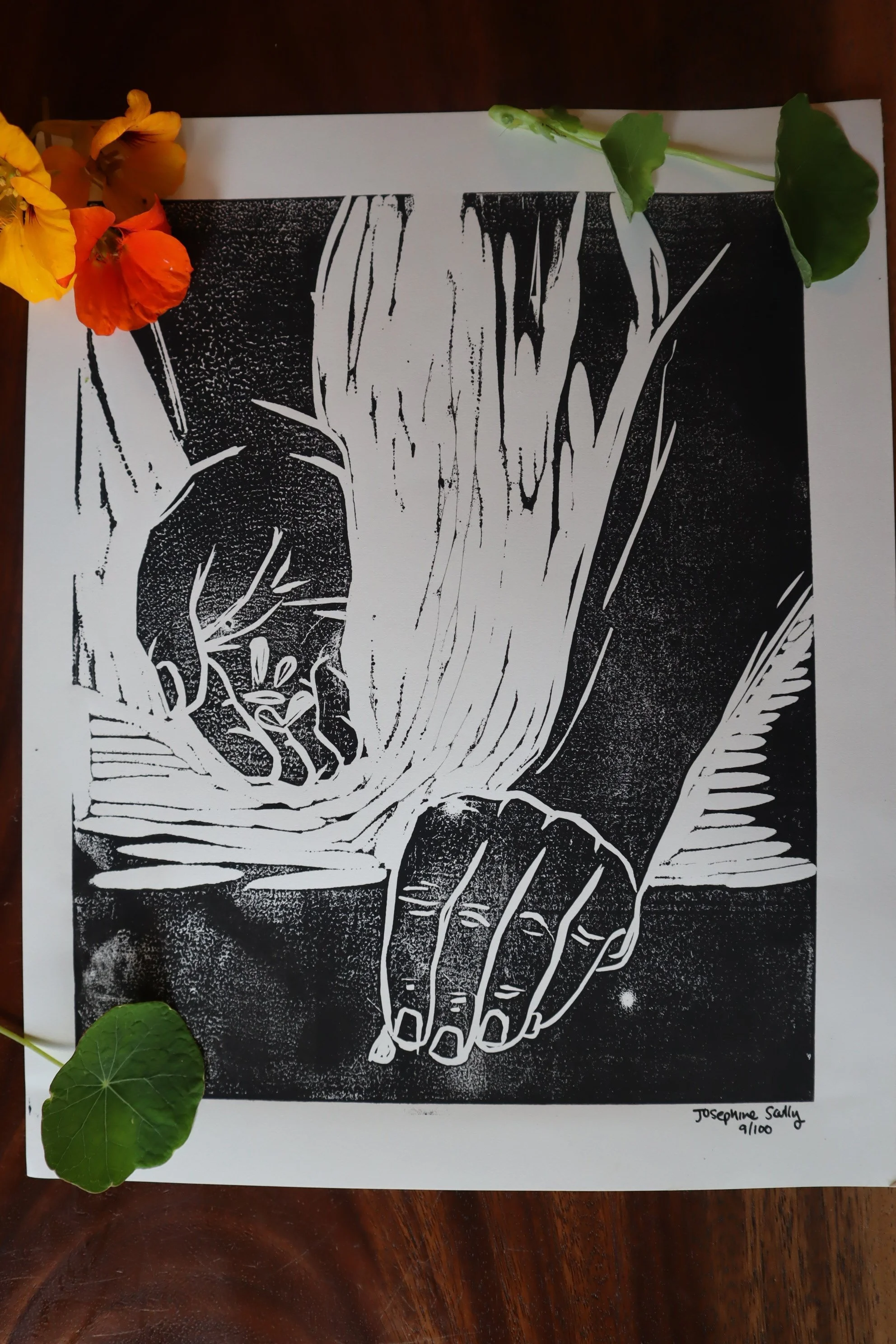 A black and white linocut print of a hand planting seeds with some yellow and orange flowers and green leaves placed on the corners, and the artist's signature and edition number at the bottom right.