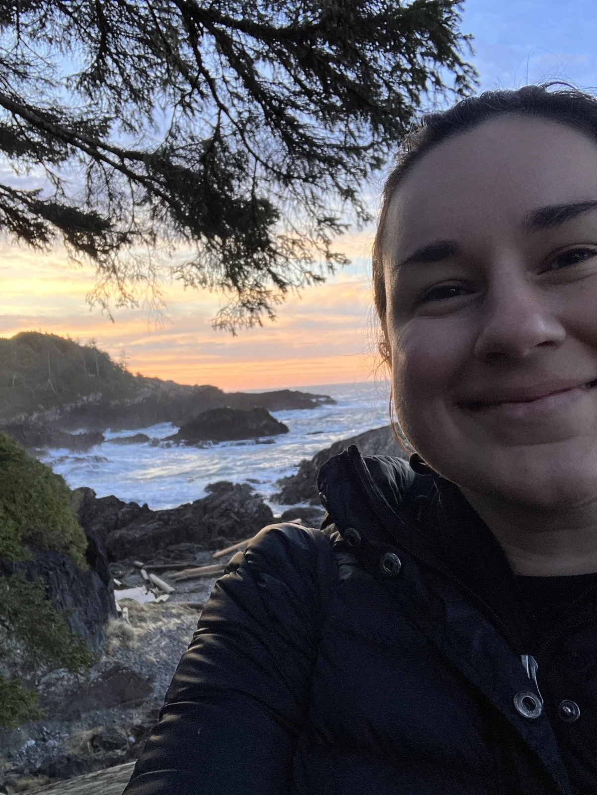 A woman taking a selfie on a rocky beach during sunset, with ocean waves behind her, trees overhead, and a colorful sky.