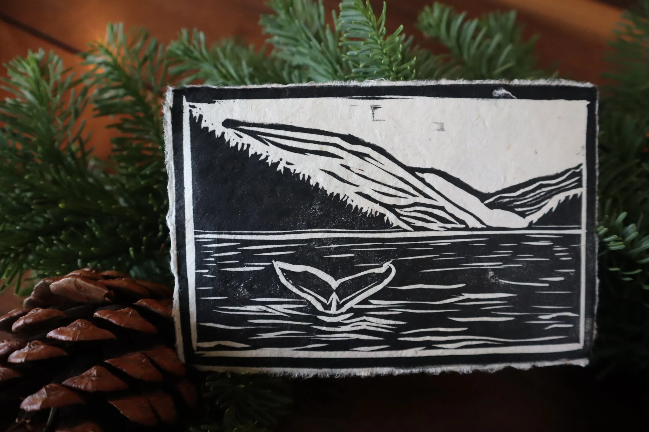 Close-up of a black and white graphic illustration depicting a mountain range and a book, set against pine branches and pinecones.