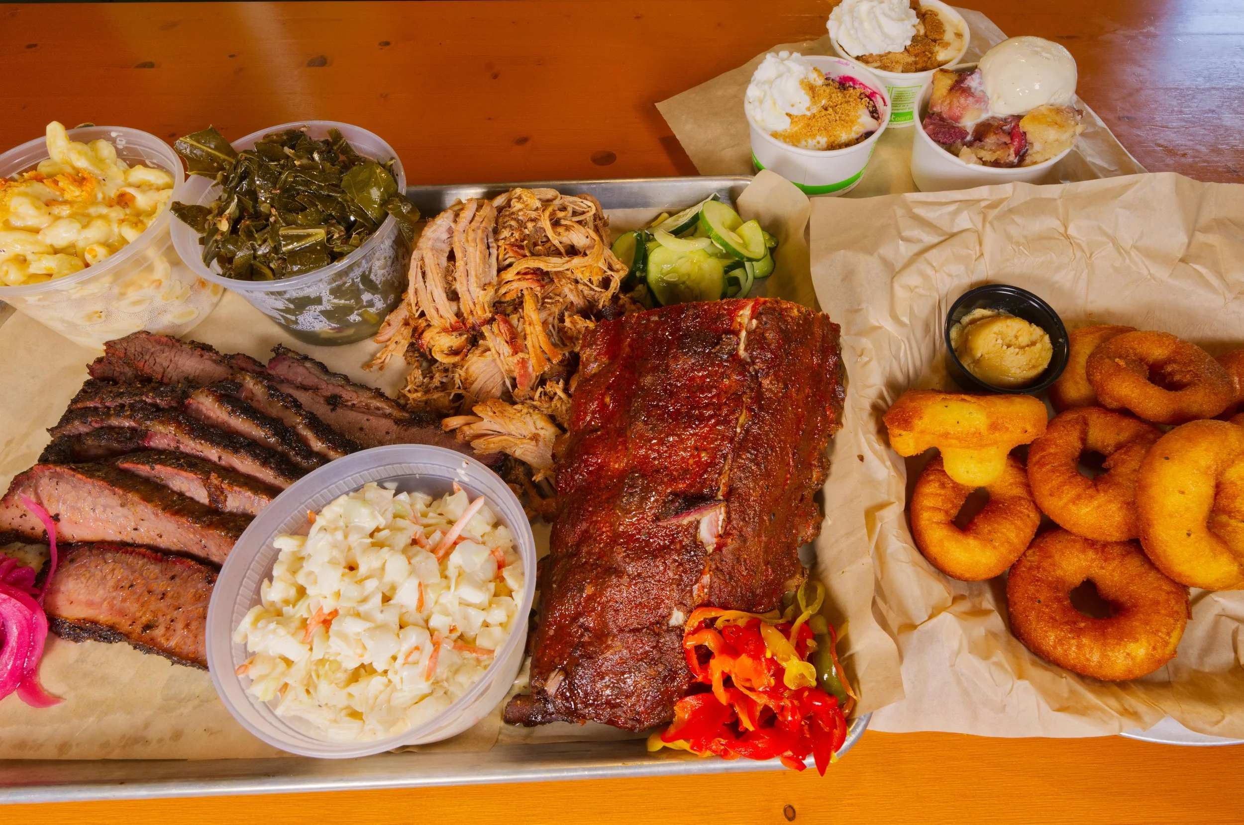 A large tray of Southern-style barbecue food spread with sides, including sliced brisket, pulled pork, baked beans, collard greens, potato salad, and onion rings, with small cups of macaroni and cheese and peach cobbler desserts.