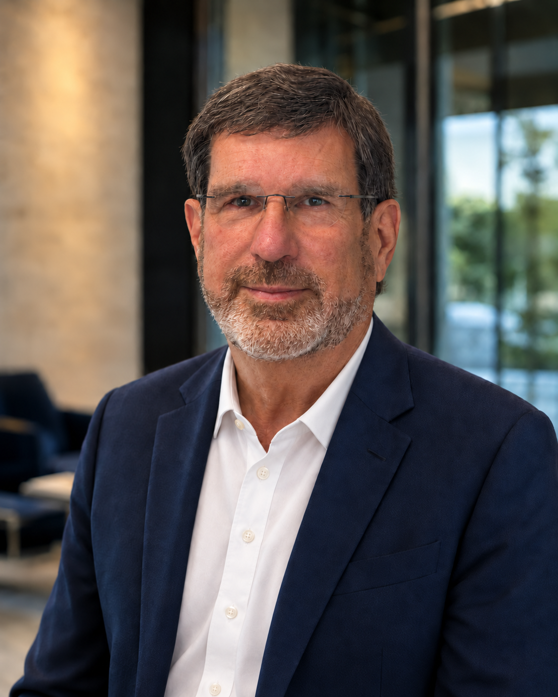 A middle-aged man with a beard and glasses wearing a navy blue suit and white shirt in an office setting.