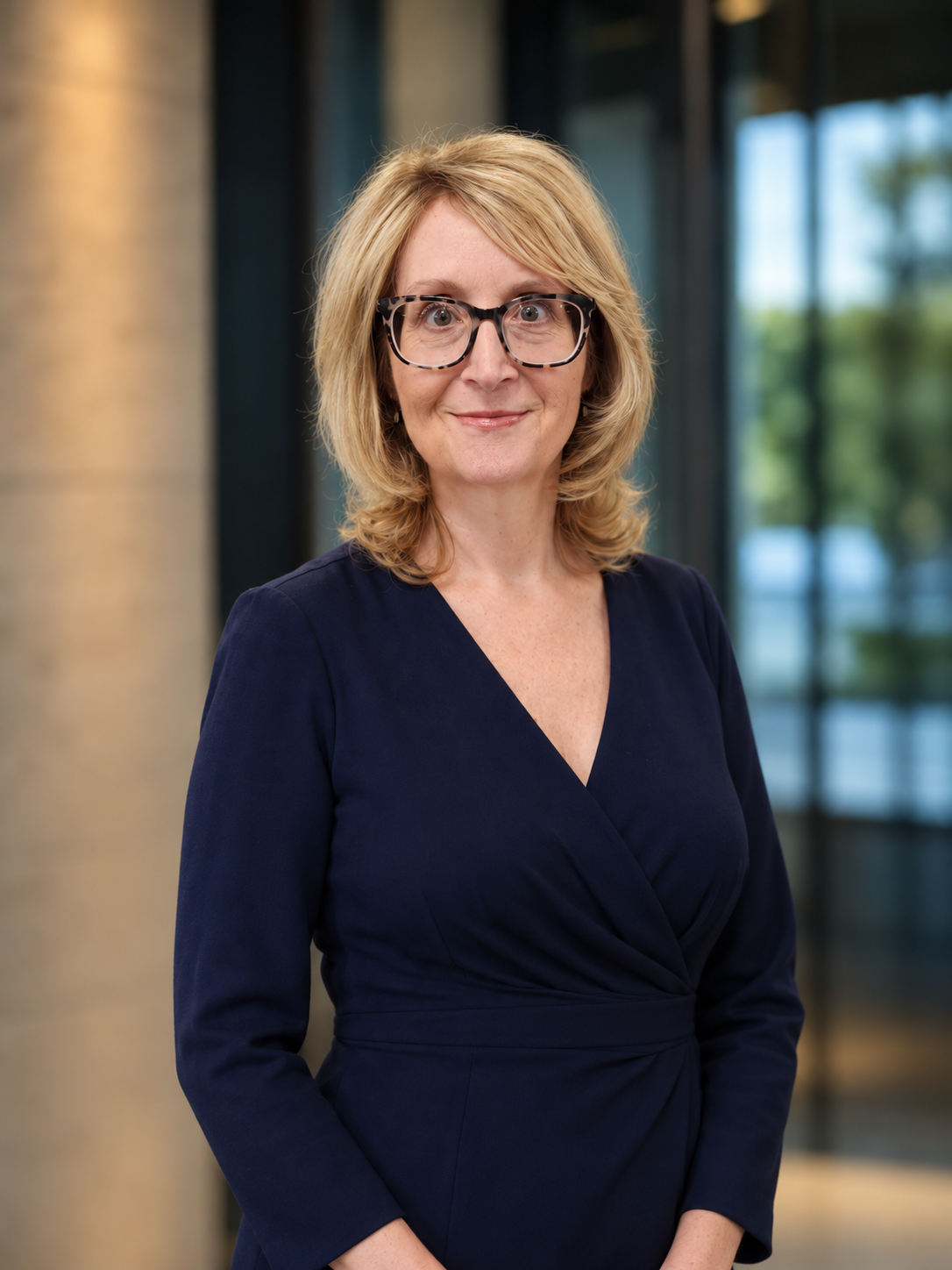 A middle-aged woman with blonde hair, wearing glasses and a dark blue dress, standing indoors with a blurred background of large windows and trees outside.
