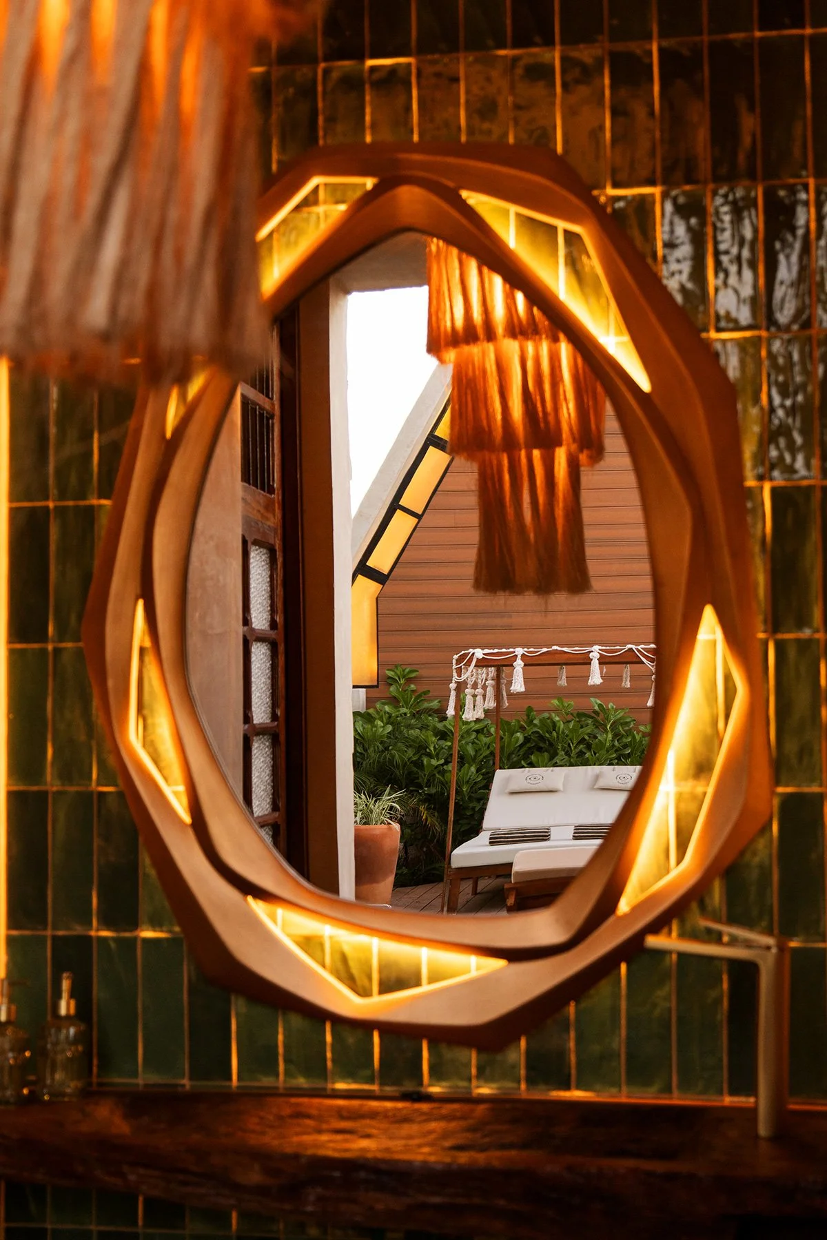 Espejo decorativo en forma de flor que refleja un patio con plantas, una cama grande con almohadas, una estructura con cortinas de fibra natural y paredes de madera.