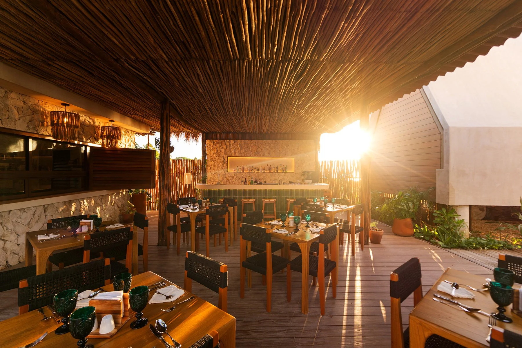 Un restaurante al aire libre con mesas de madera y sillas negras, decorado con plantas y una barra de bar, durante el atardecer con luz cálida que entra por el lado derecho.