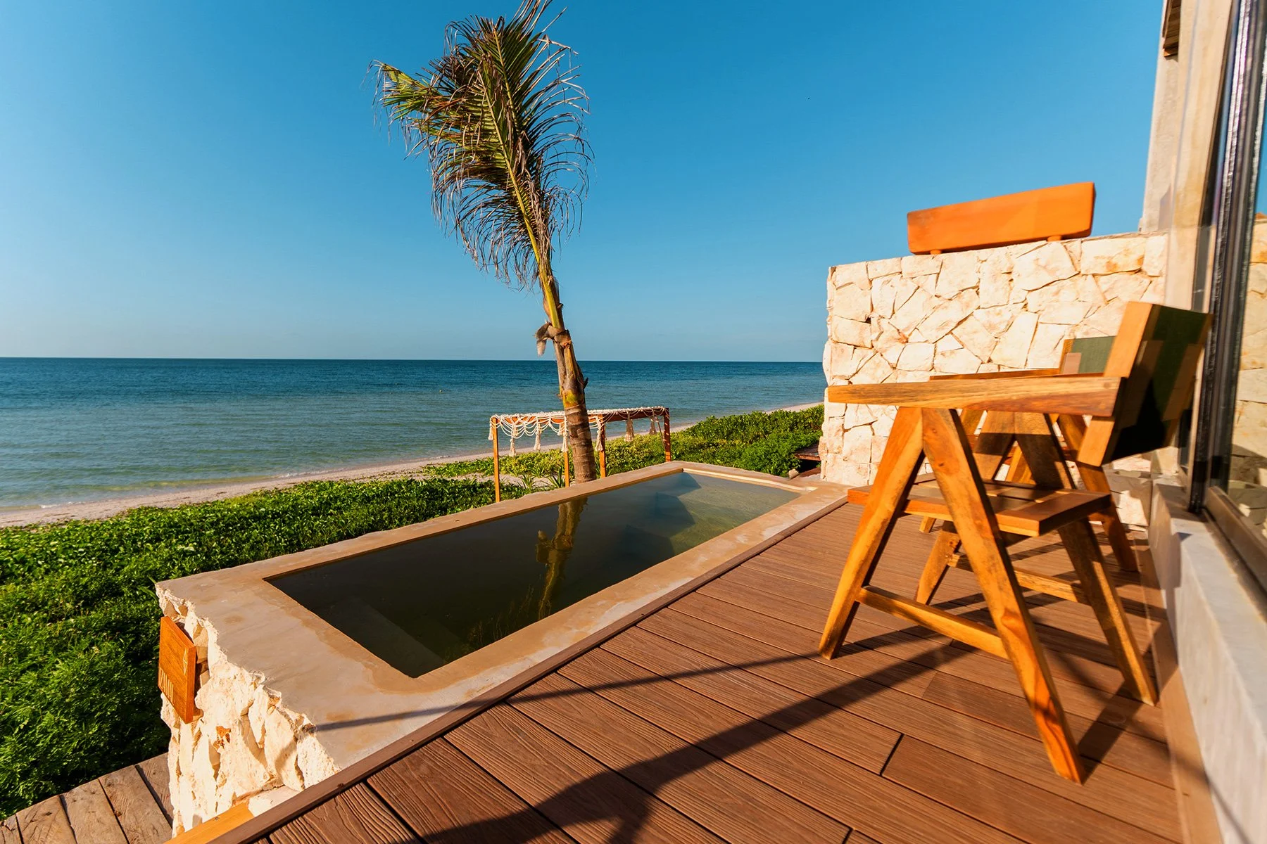 Vista de una terraza junto a la playa con sillón de madera, palmera, piscina pequeña y vista al mar.