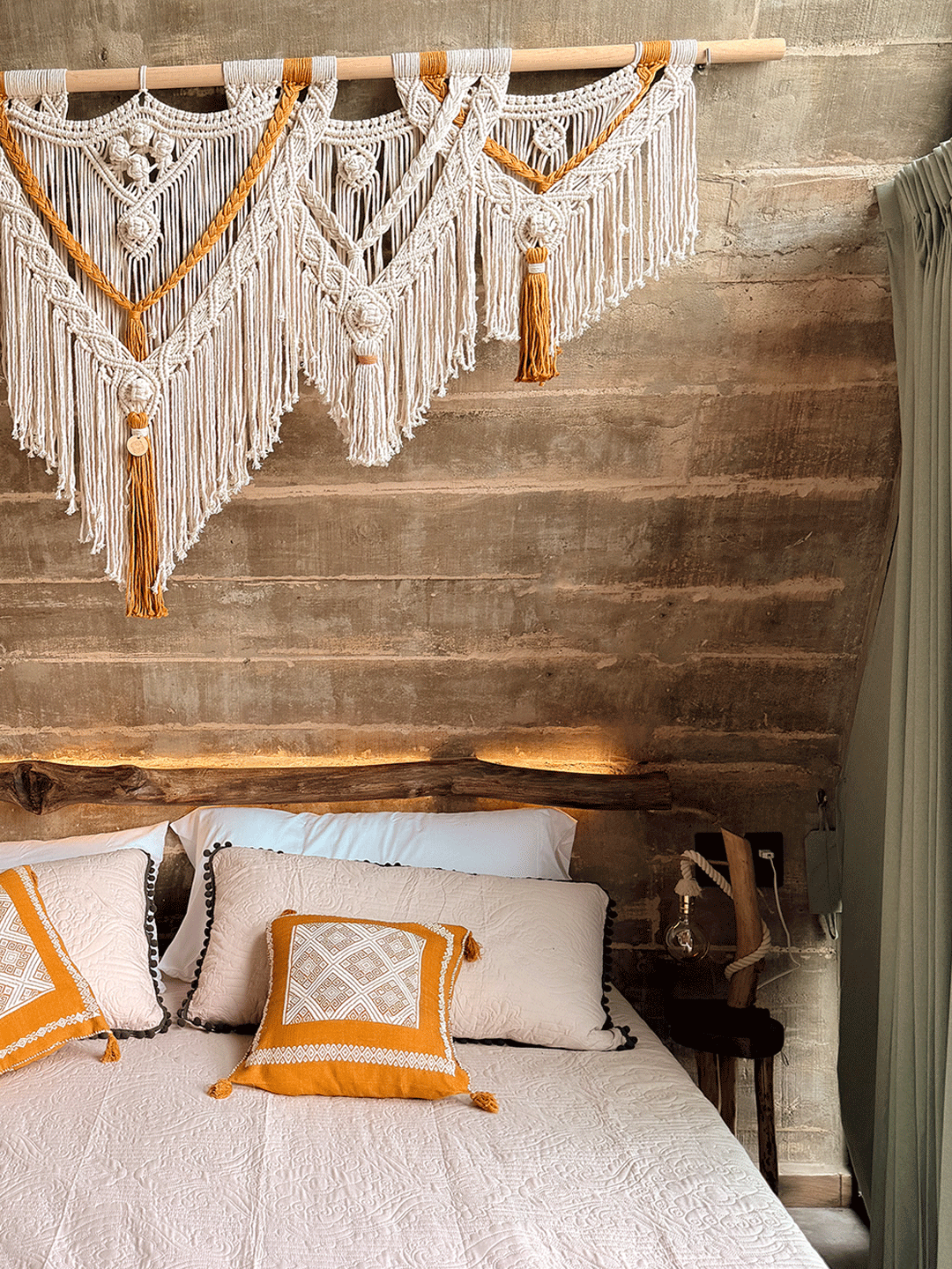 Cama con almohadas blancas y detalles en color naranja, tapiz de macramé decorativo en la pared y cortinas verdes a la derecha.