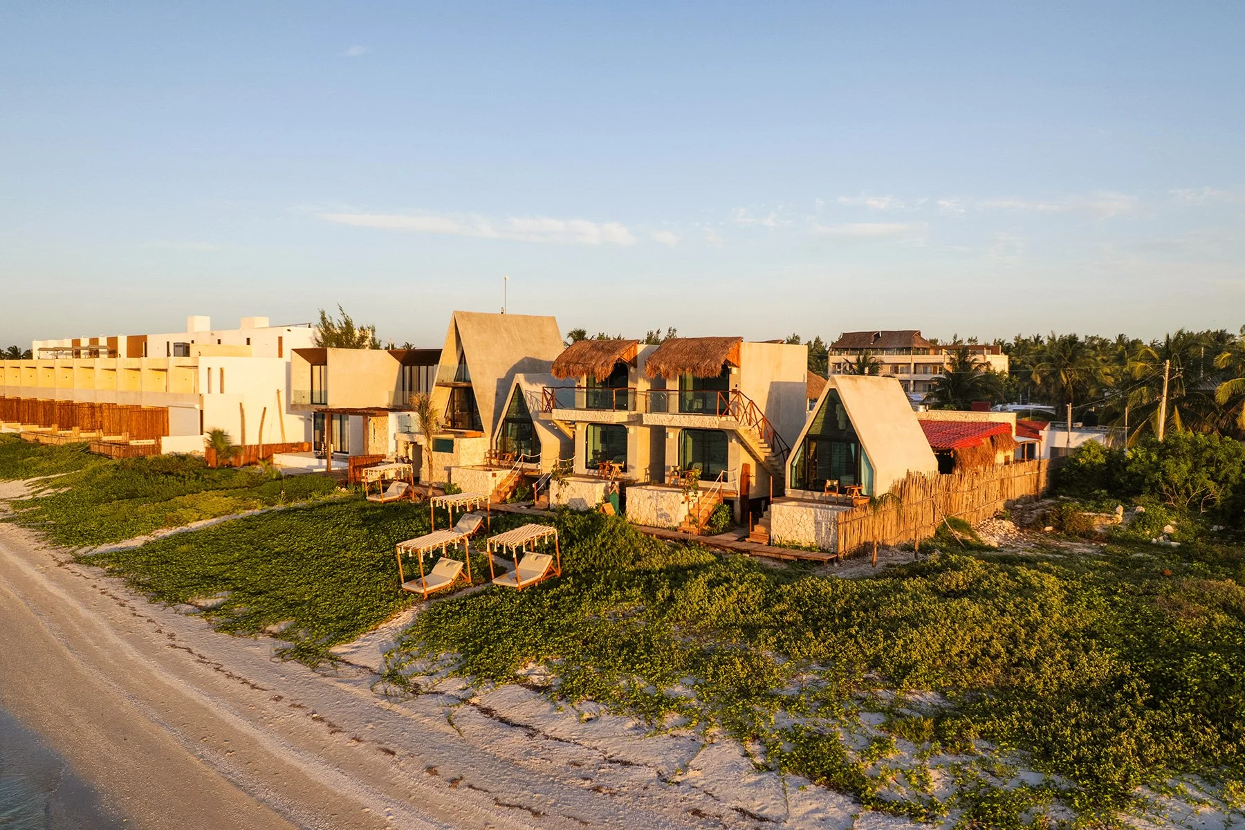 Conjunto de cabañas modernas en la playa durante el atardecer, con áreas de descanso y vegetación alrededor.