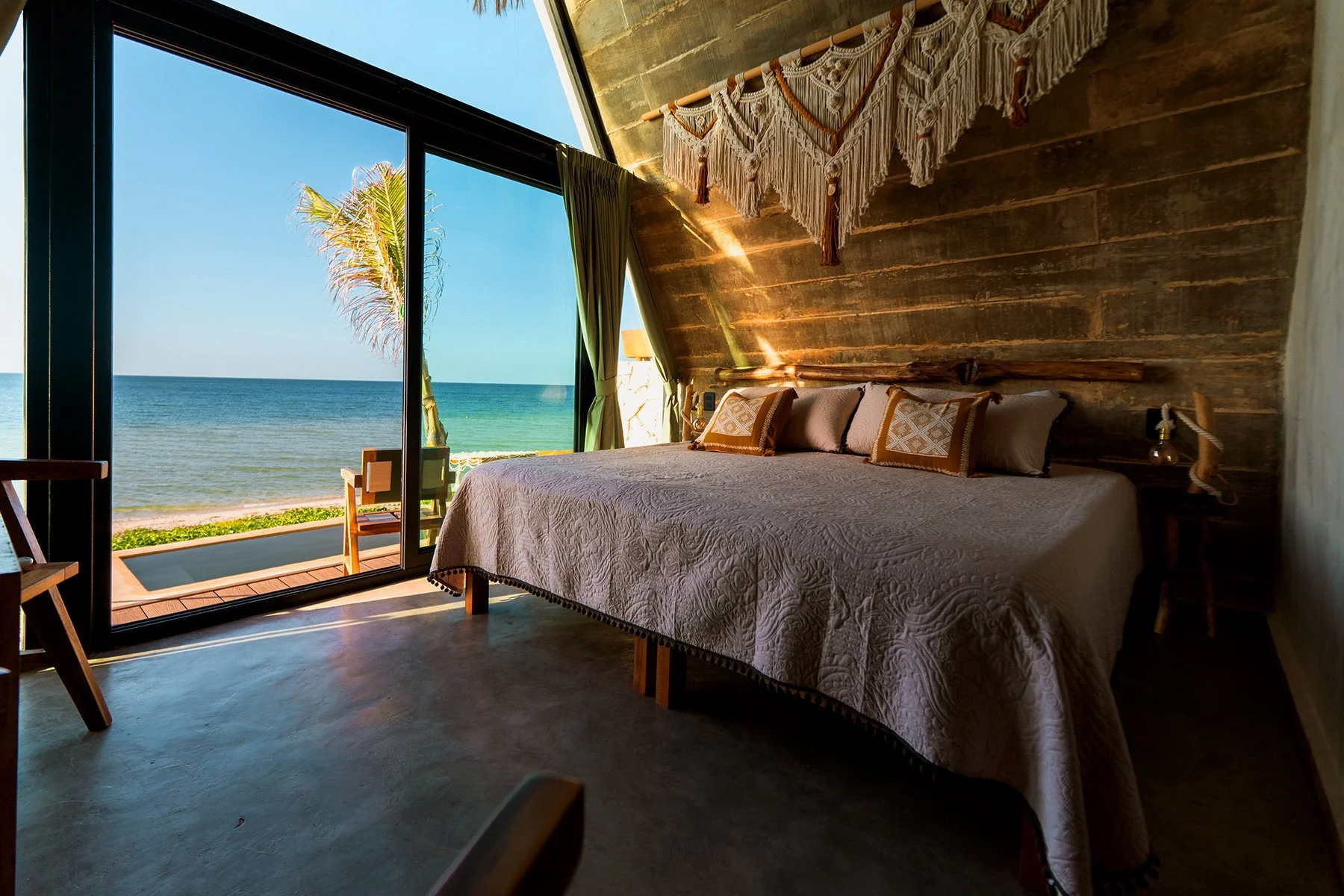 Habitación con vista al mar, cama con ropa de cama blanco, almohadas decorativas, lámpara de mesa, cortinas verdes, decoración bohemia, pared de madera, ventana grande con vista a la playa y palmeras.
