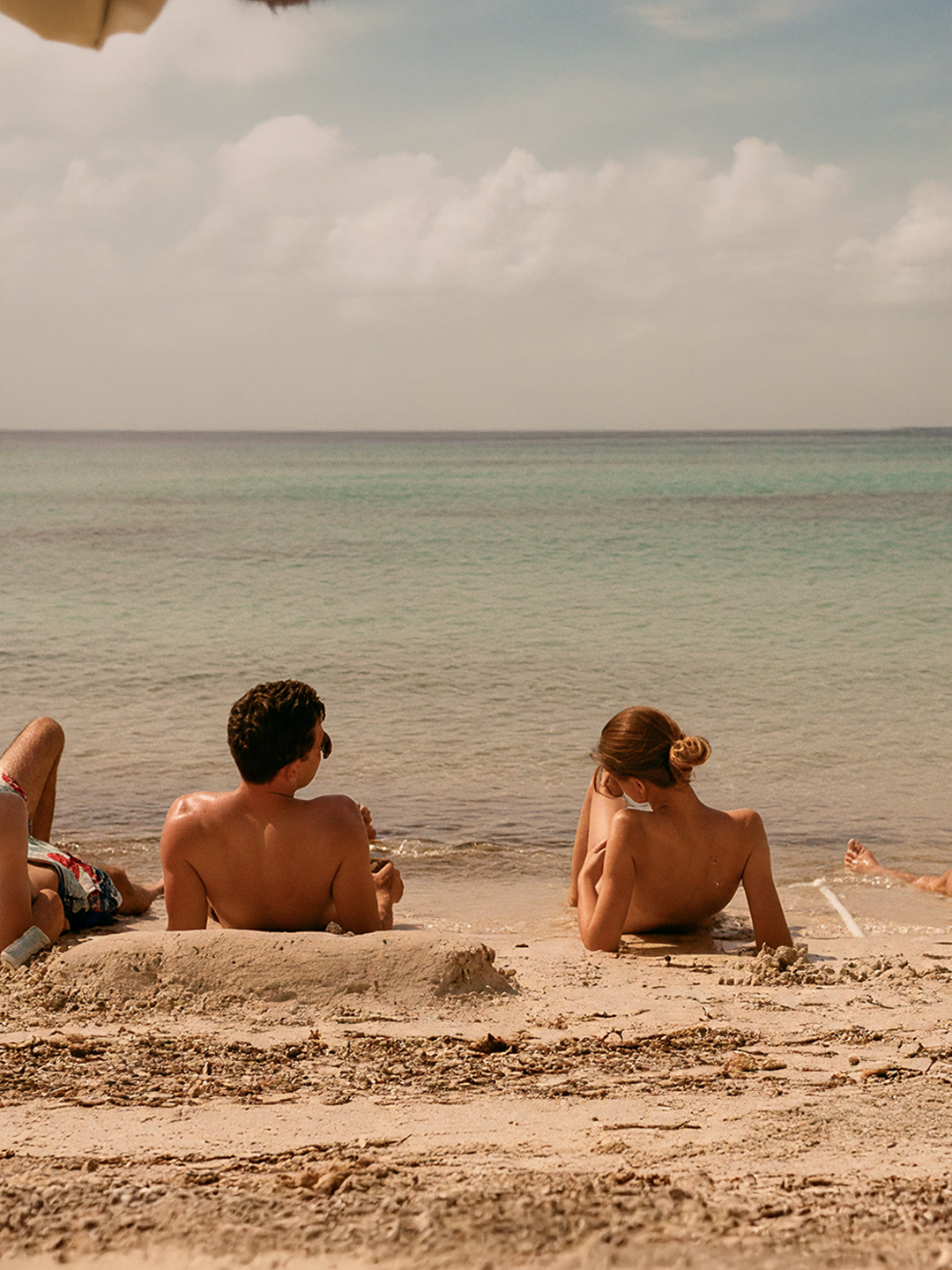 Pareja de dos personas, un hombre y una mujer, sentados en la playa en la arena, mirando hacia el mar con poca gente alrededor, en un día soleado con cielo parcialmente nublado.