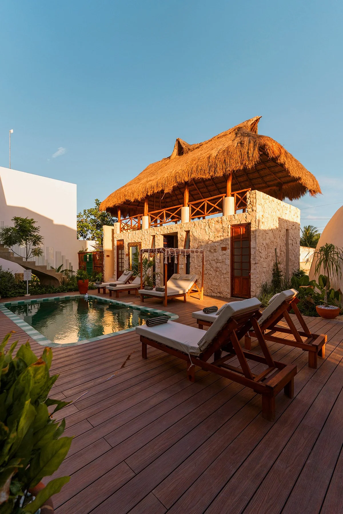 Casa de estilo rústico con techo de palma, balcón amplio, piscina, sillones con sombrillas, y jardín con plantas tropicales, durante el atardecer.