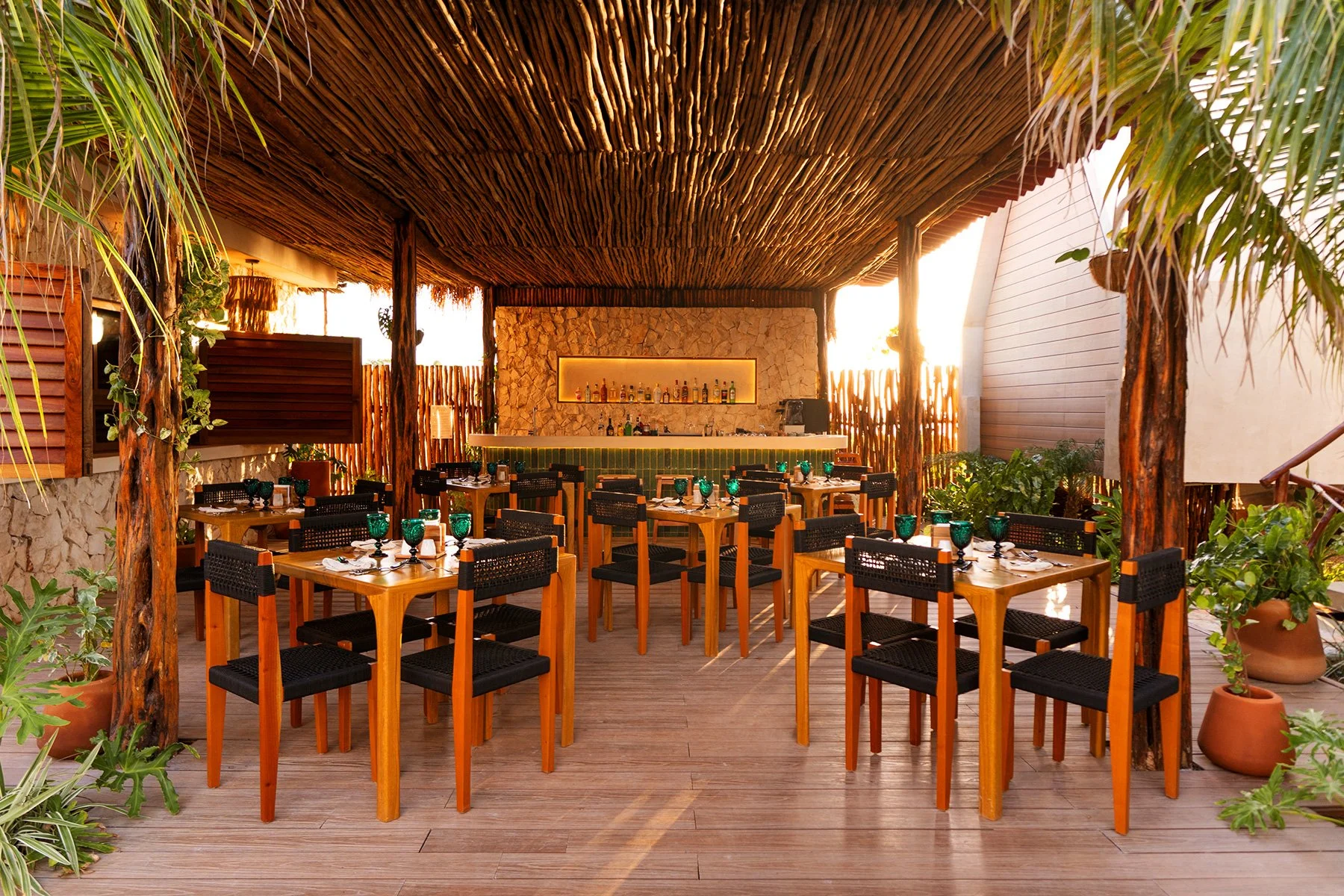 Área al aire libre con mesas de madera y sillas negras, decorada con plantas en macetas y una estructura de techo de madera y cañas. En el fondo, una barra de bar con lámpara de luz cálida, rodeada de paredes de piedra y madera, con vegetación en los