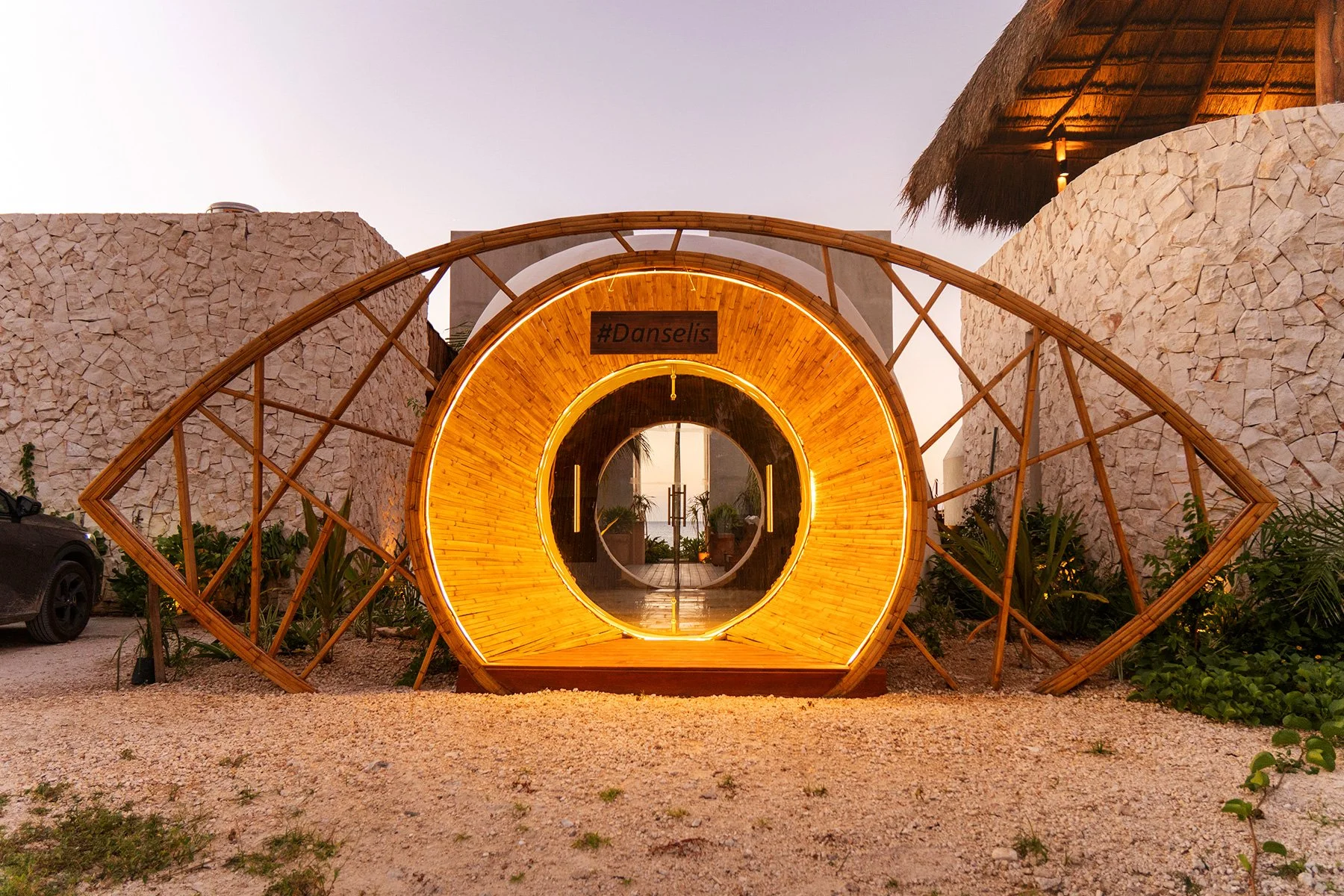 Entrada decorativa de madera con luces de neón y una señal que dice #Danselis. El entorno incluye paredes de piedra y plantas, con una estructura que combina elementos naturales y modernos.