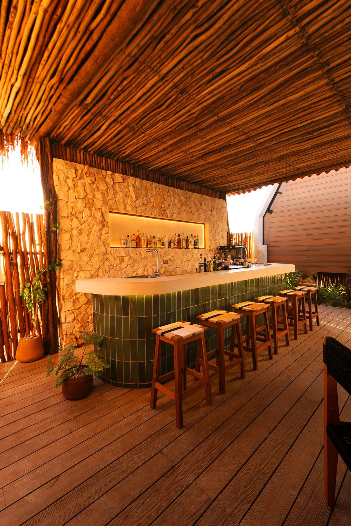 Bar con barra de cerámica verde y taburetes de madera, paredes de piedra y cerca de madera, techo de madera y plantas decorativas.