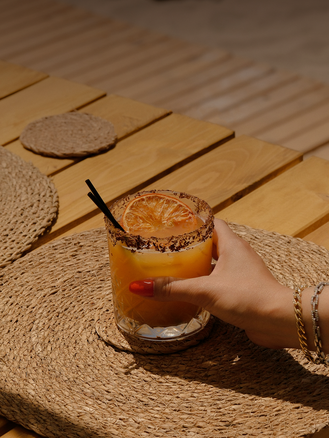 Persona sosteniendo una bebida con rodaja de naranja, en un vaso con borde escarchado, sobre una mesa de madera con accesorios de ratán.