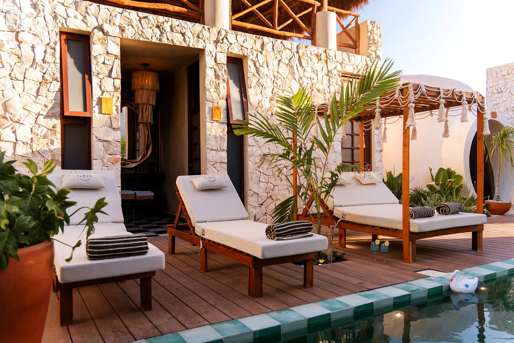 Área de piscina con tumbonas blancas y cojines, entre plantas tropicales y un techo de cañas, en un resort elegante.
