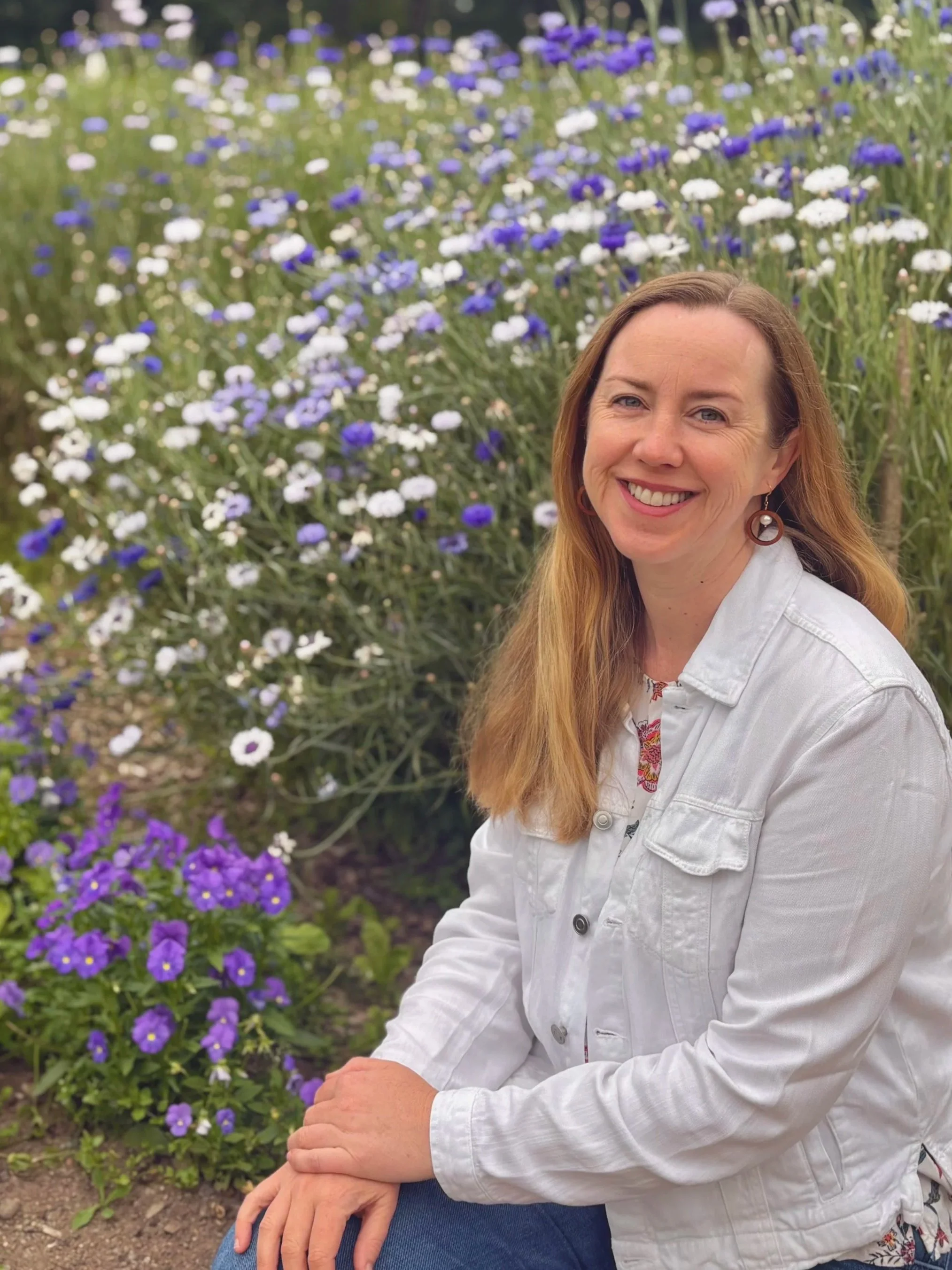A woman sitting outdoors in front of a flower bed with purple and white flowers, smiling and wearing a white jacket and earrings.