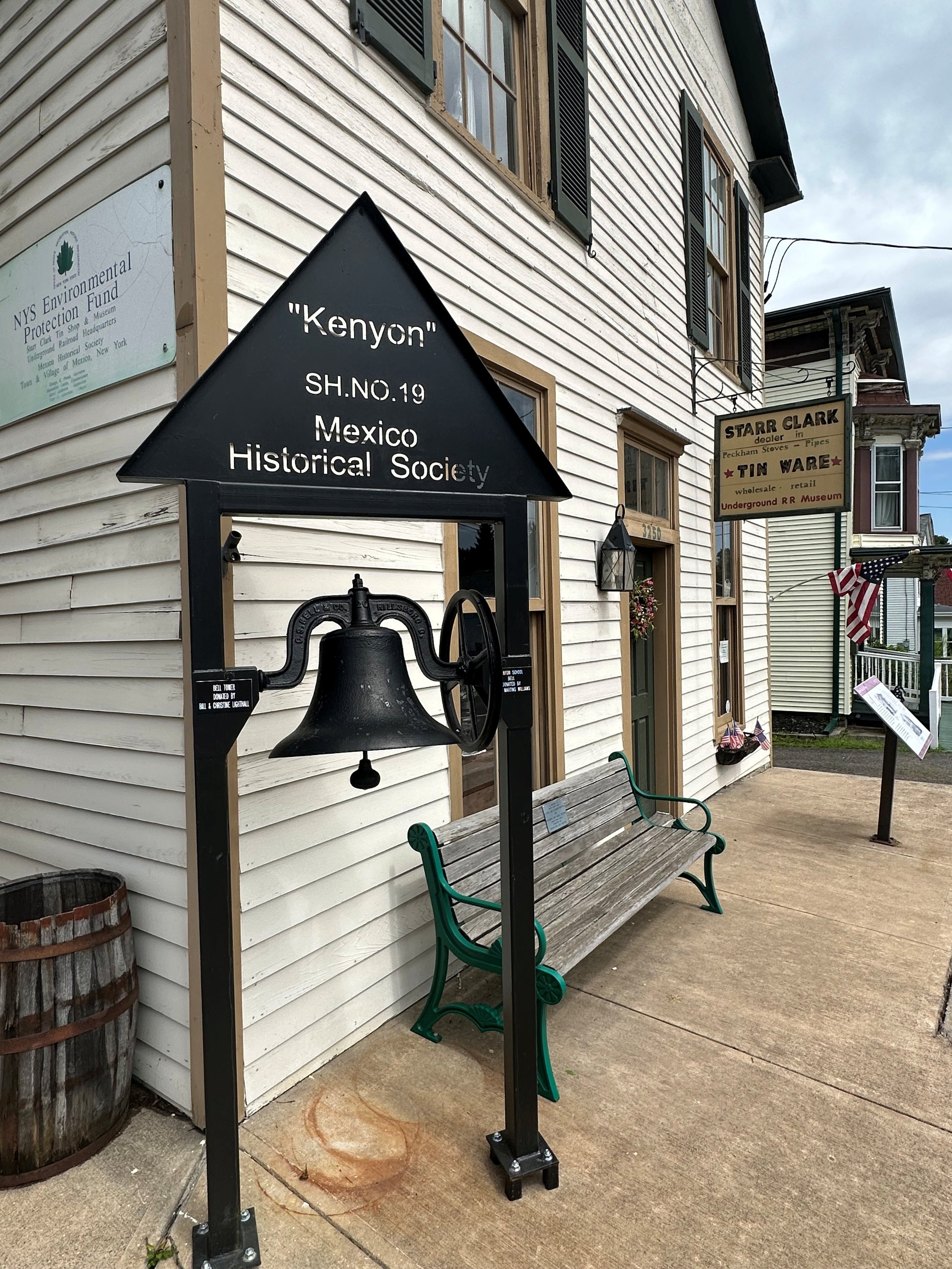 A historic building with white wooden siding, black window shutters, a bell, a bench, and American flags outside.