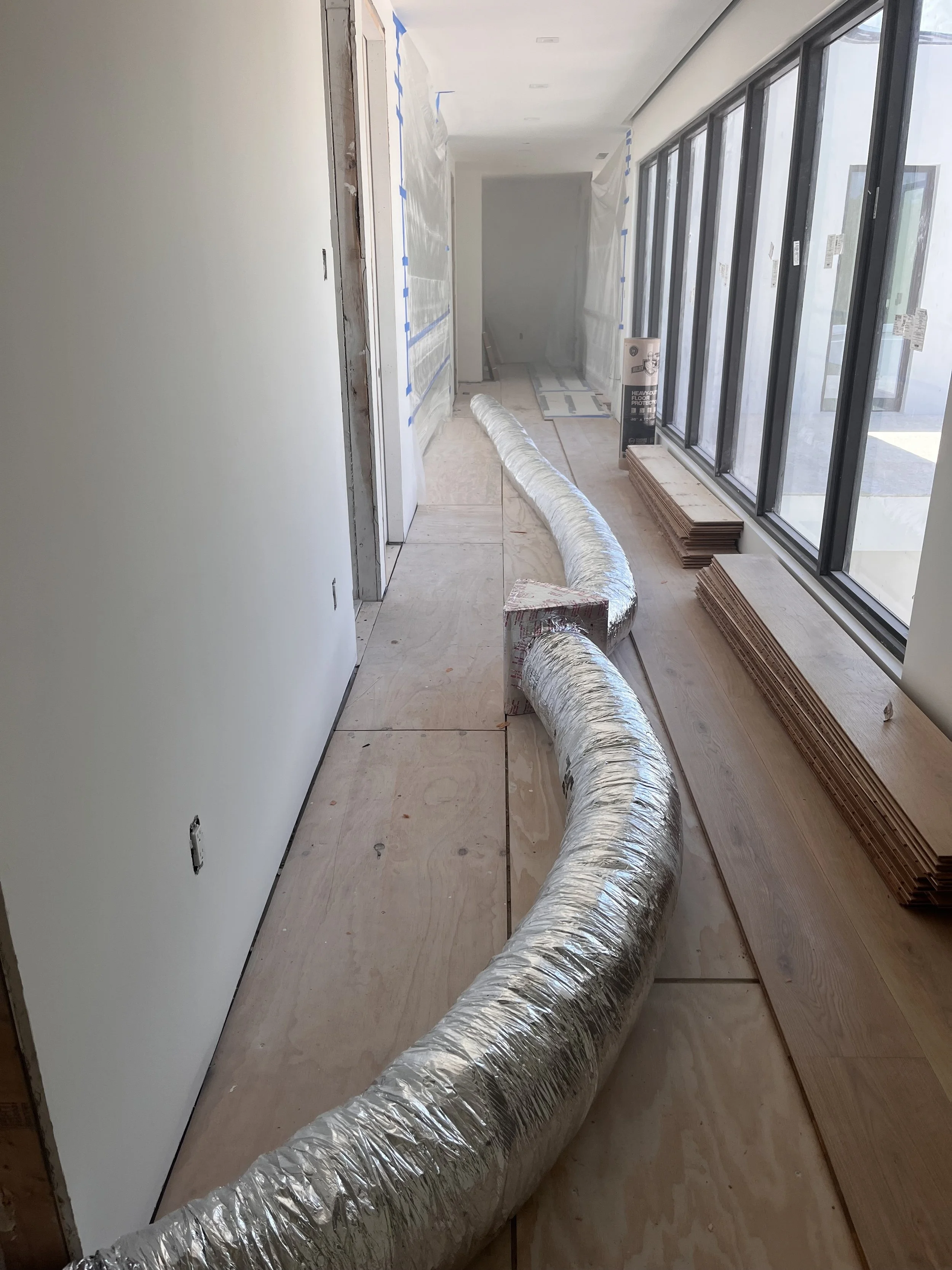 A long, silver, flexible duct runs along the floor of an indoor construction site, with building materials and windows on one side and a partially covered doorway on the other.