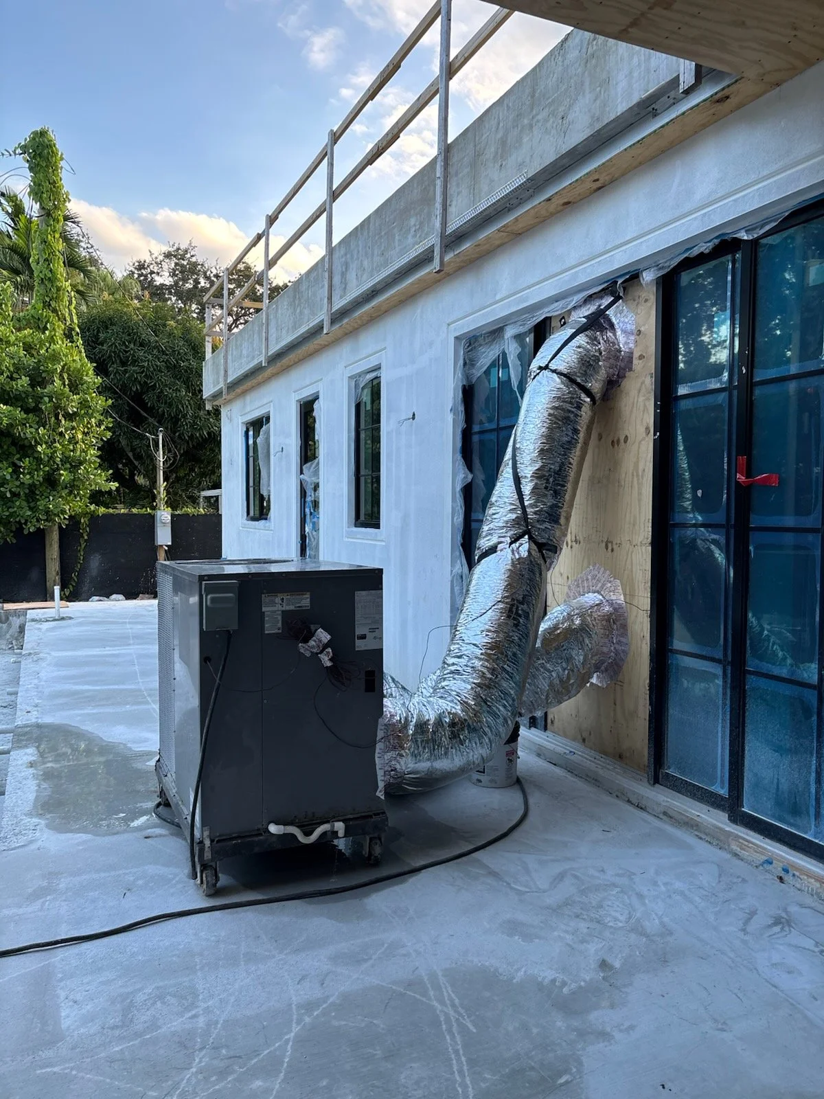 Construction site of a building with window frames, ventilation ducts, and insulation work in progress, outdoor setting with trees and partly cloudy sky.