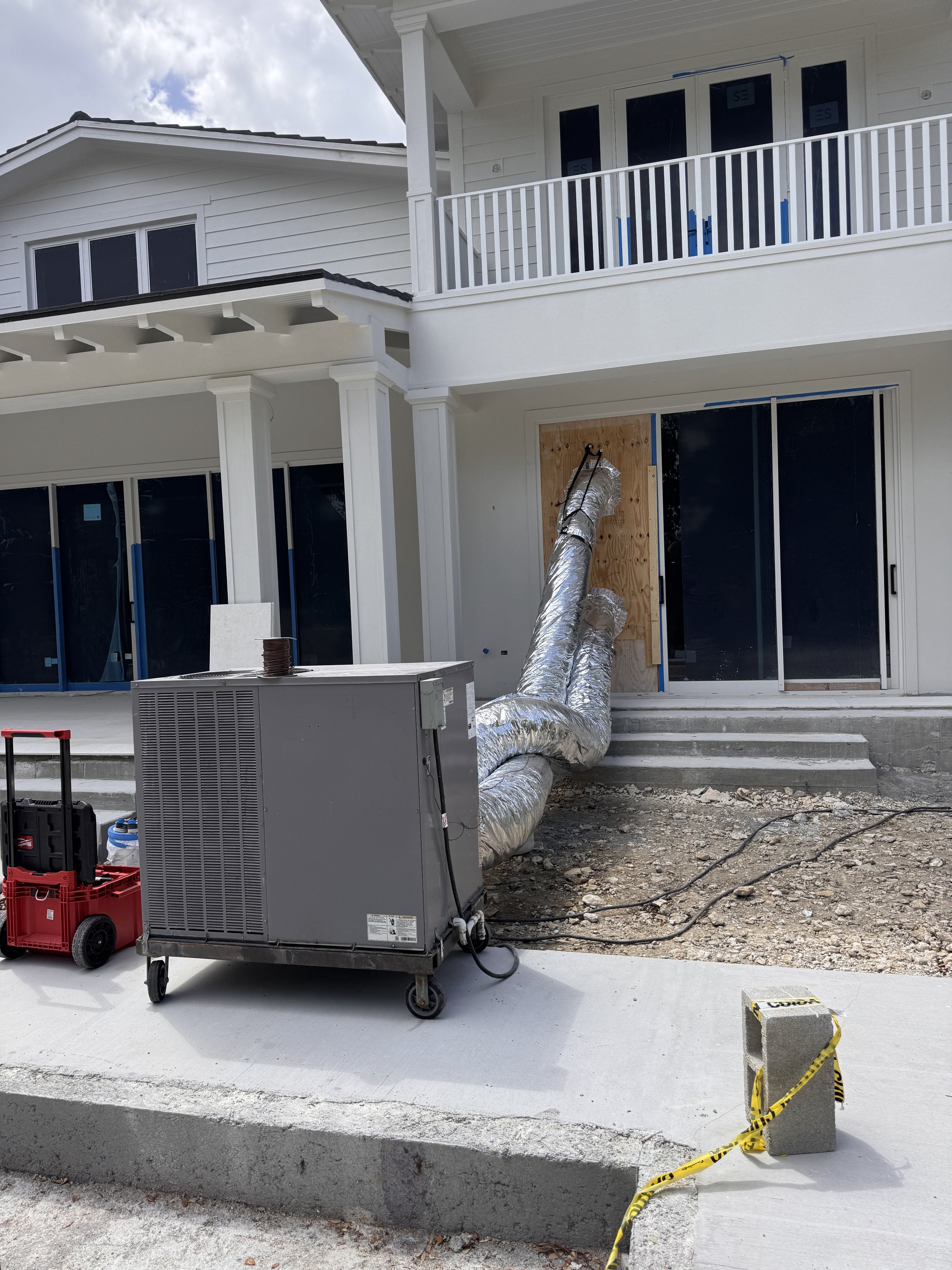 Construction site of a modern house with white exterior, large glass doors, and sliding windows. Visible HVAC equipment, ductwork wrapped in insulation, and construction tools are present. The area is partially cordoned off with caution tape.