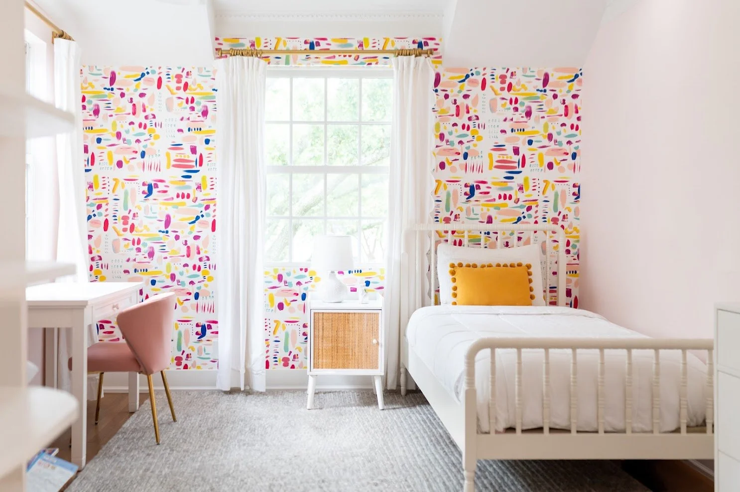 Bright bedroom with colorful abstract wallpaper behind a white bed with a yellow pillow, a white bedside table with a white lamp, a pink chair with gold legs, and a large window with white curtains revealing green trees outside.